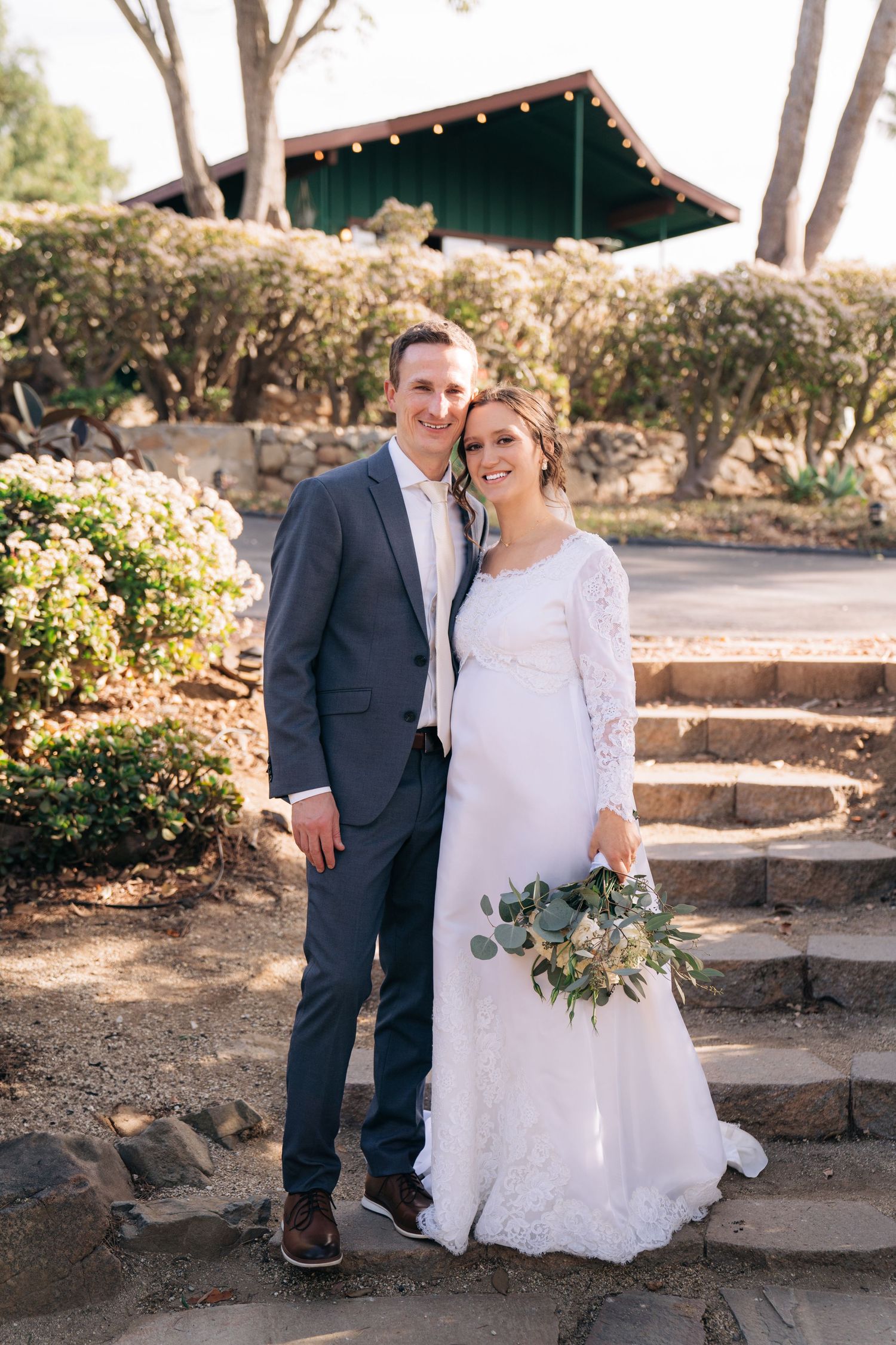 A couple stands together on stone steps in a garden setting, with the bride wearing a lace gown and holding a eucalyptus bouquet.
