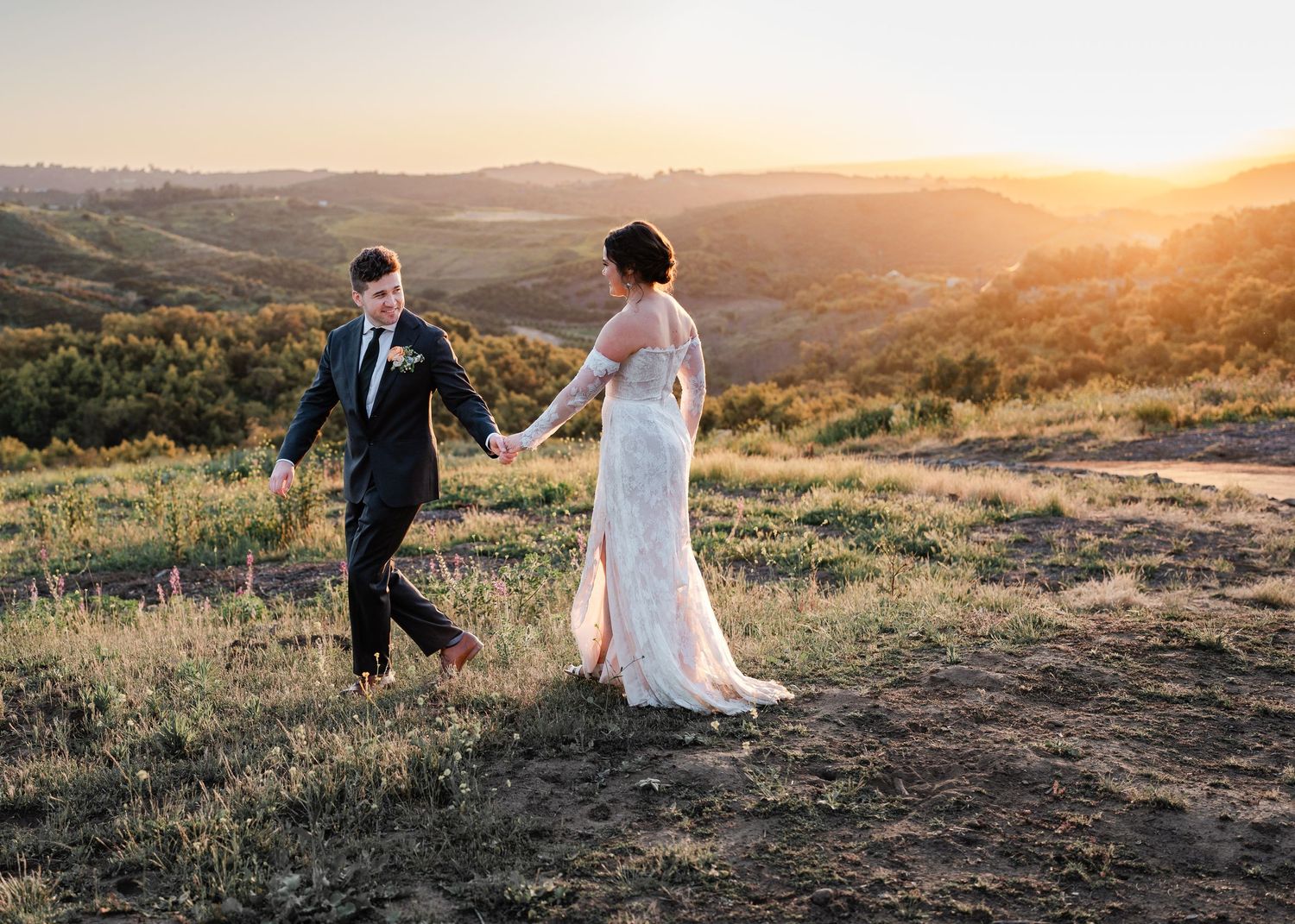 A couple in wedding attire walking hand in hand on a hilltop at golden sunset in a scenic countryside location.