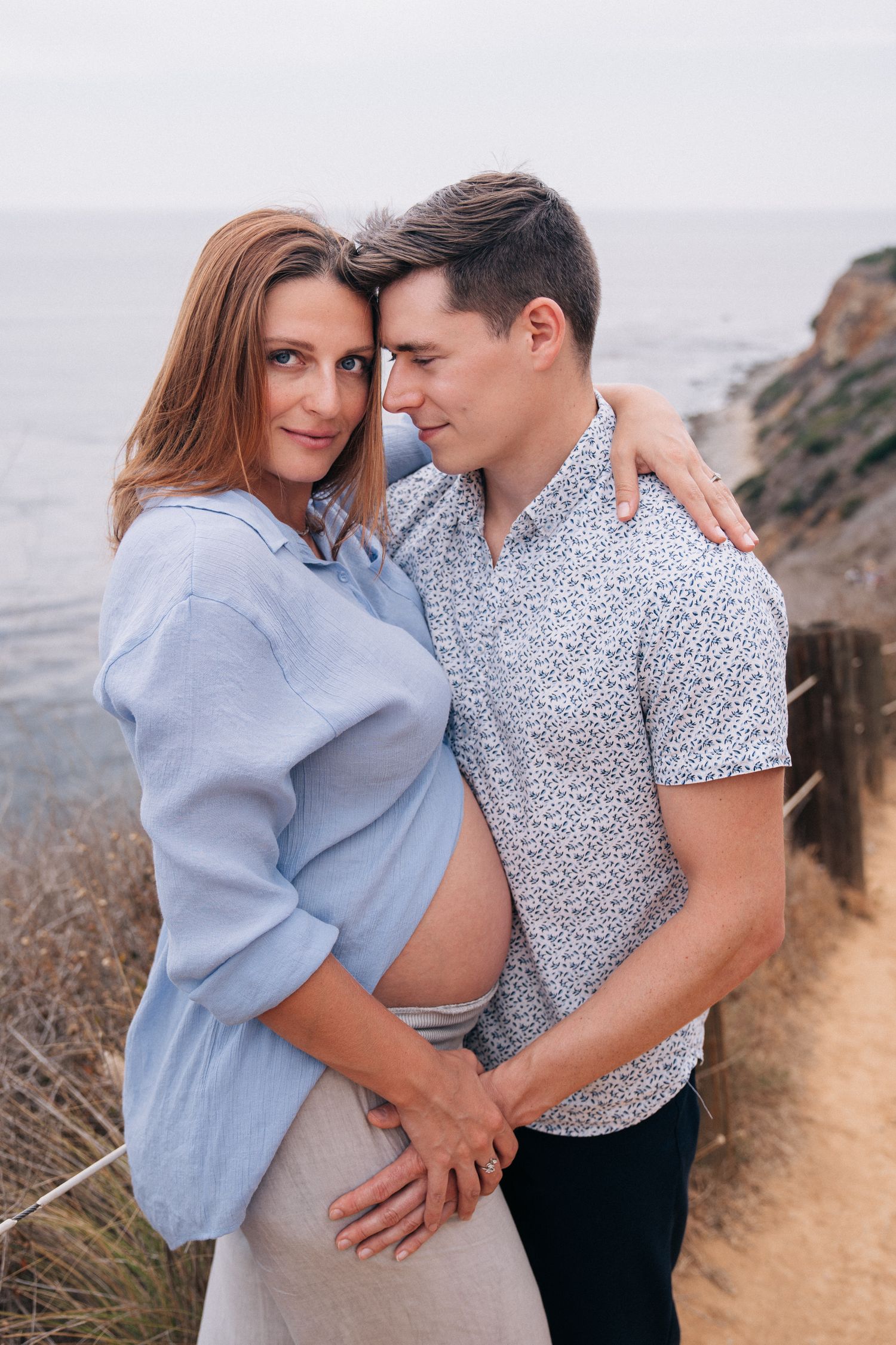 A couple embraces on a coastal cliff path overlooking the ocean on a misty day.