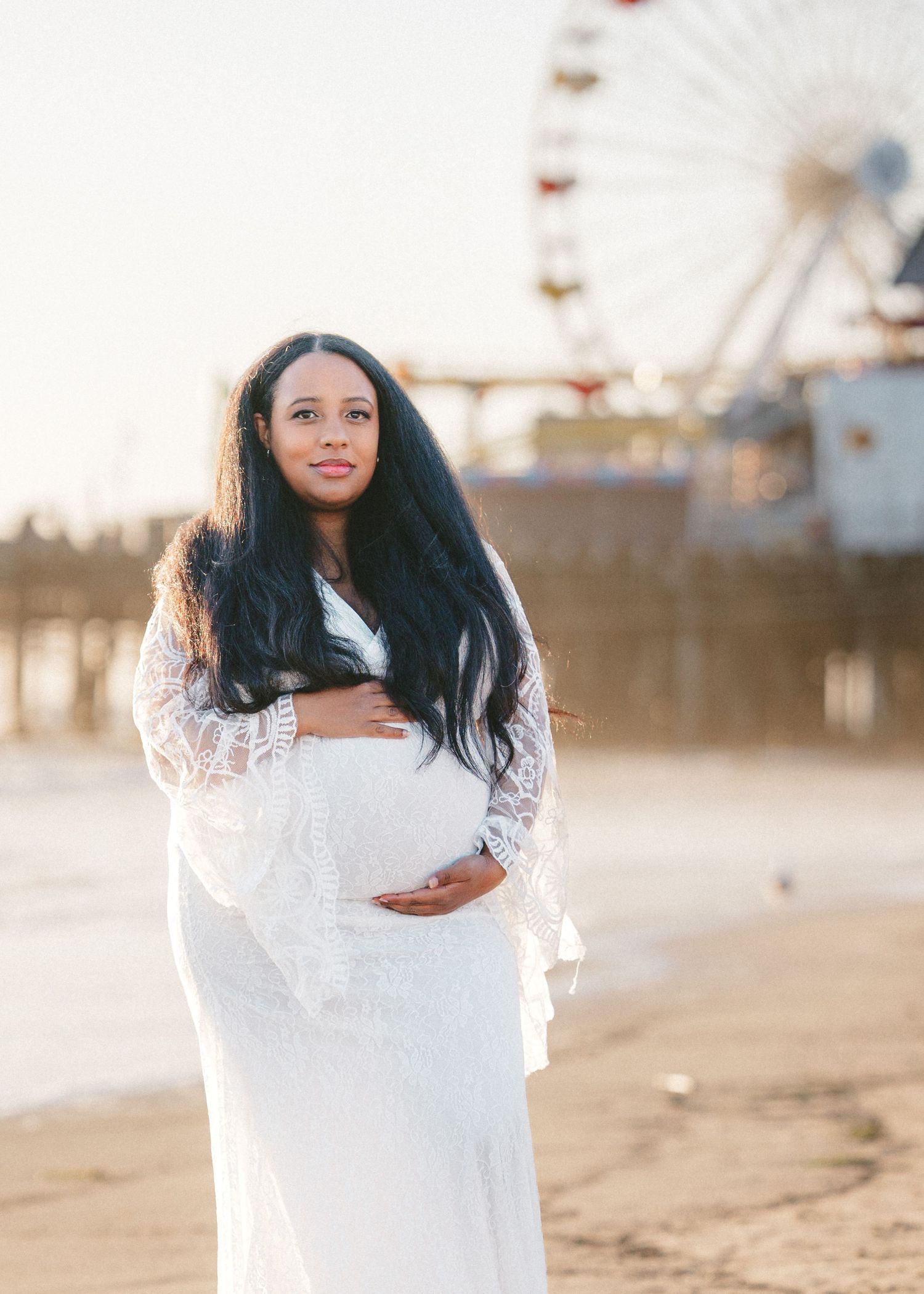 Woman in white lace dress poses on sandy beach at sunset in series of romantic maternity photos.
