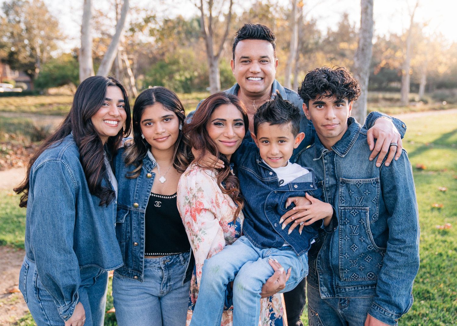 A group of five people wearing denim jackets pose together outdoors in a park during autumn.