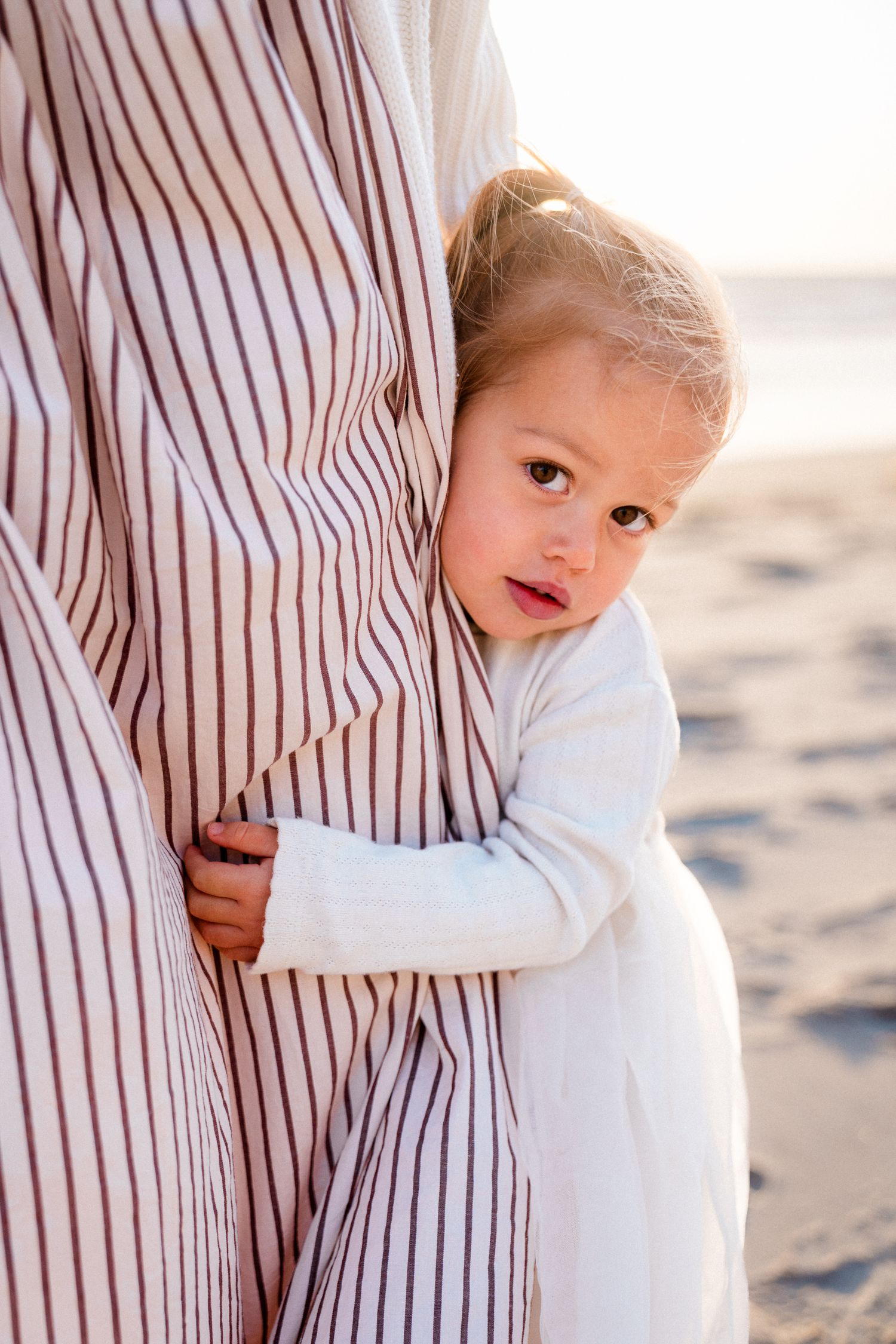A child in white clothing hugs a person wearing striped clothing while standing on a sandy beach at sunset.