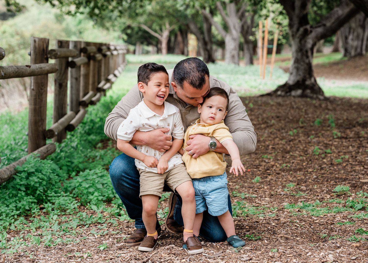 Three people sharing a tender outdoor moment together on a wooded path with wooden fencing and green foliage.