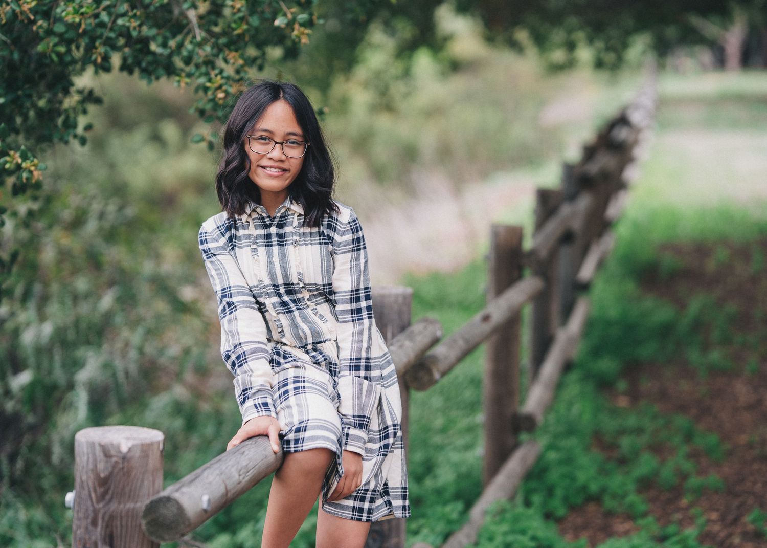 Someone wearing glasses and a plaid dress leans against a wooden fence post in a natural outdoor setting.