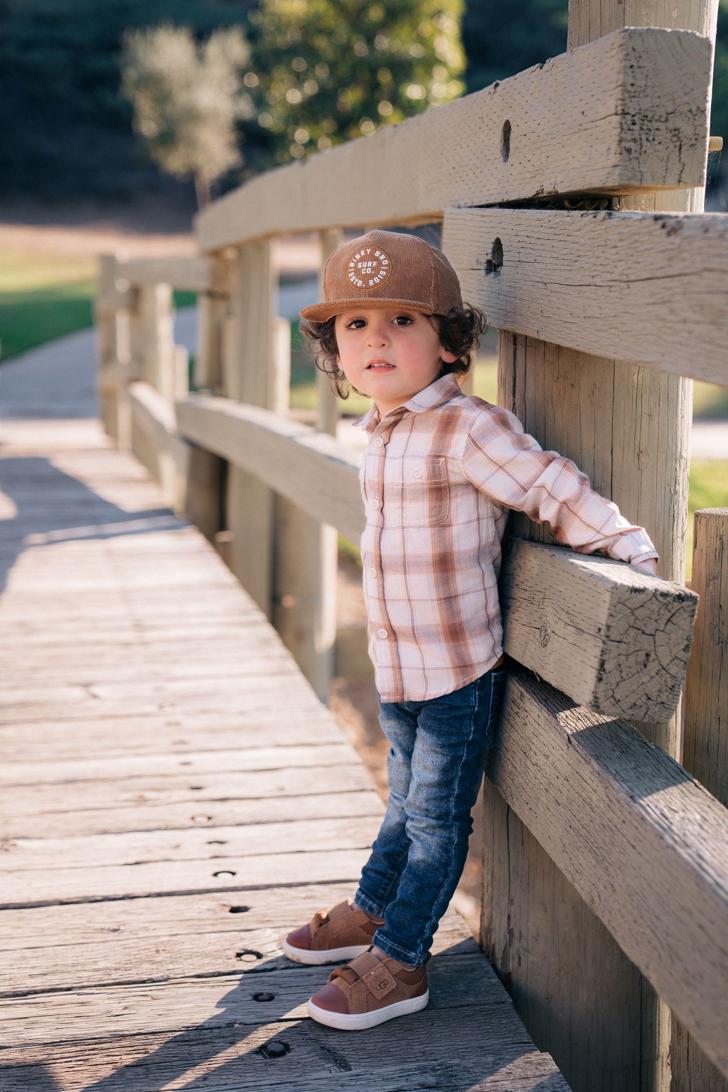 A young child in plaid shirt and baseball cap leans against a wooden railing on a sunlit bridge deck.