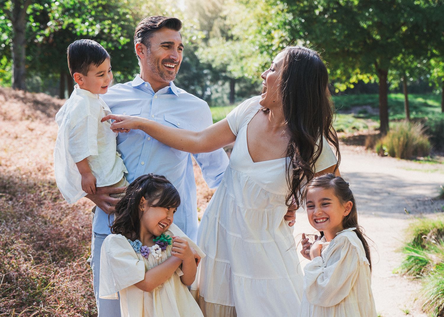 A happy family wearing white outfits laughs and plays together during an outdoor photo session in a park.