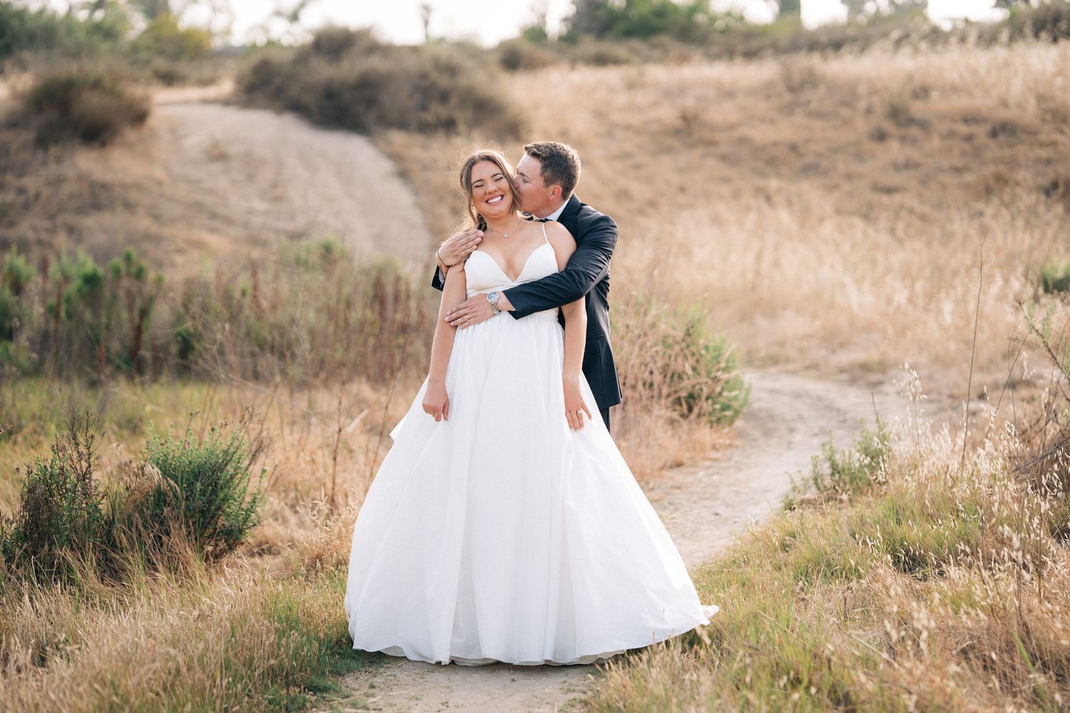 Joyful couple shares romantic moments in white wedding attire against a rustic outdoor backdrop with golden grass.