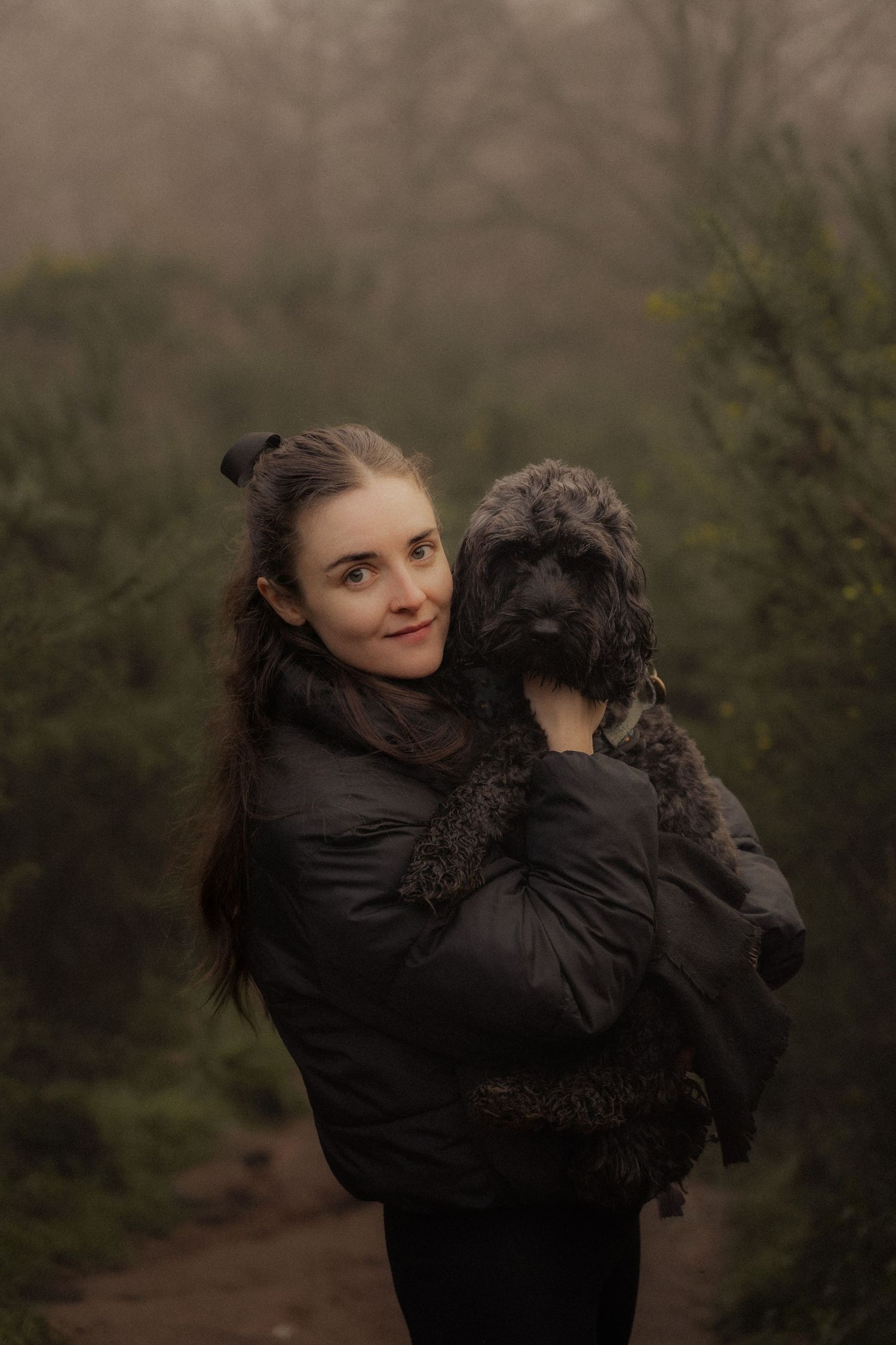 Person cuddling with large black furry dog outdoors on foggy day.