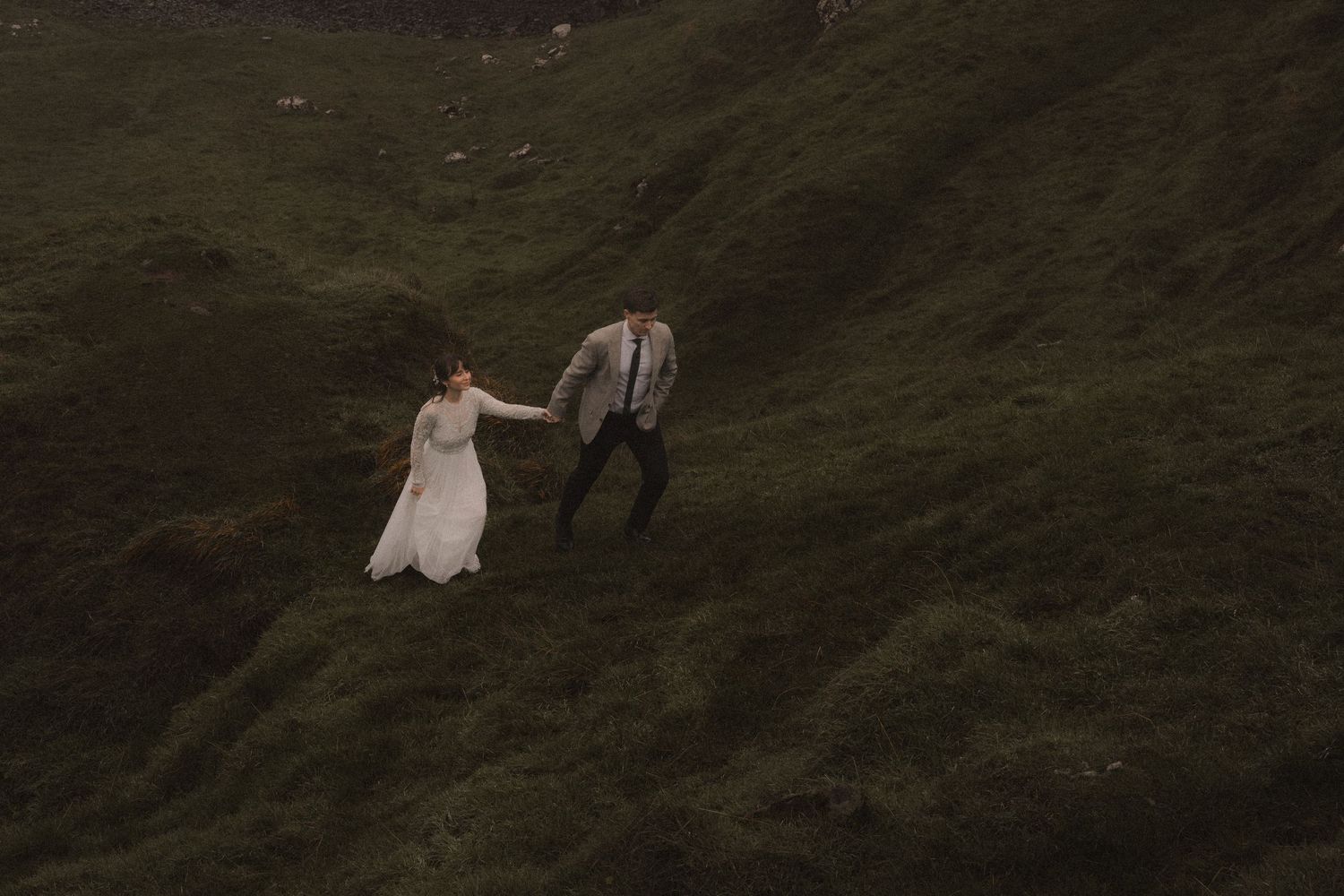 A couple in wedding attire walking hand in hand on a steep grassy hillside.