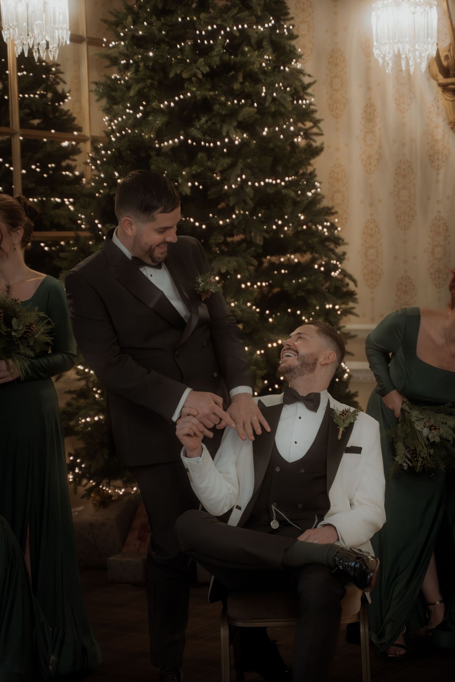 Two people share a tender moment while laughing together in front of a lit Christmas tree at a formal holiday gathering.