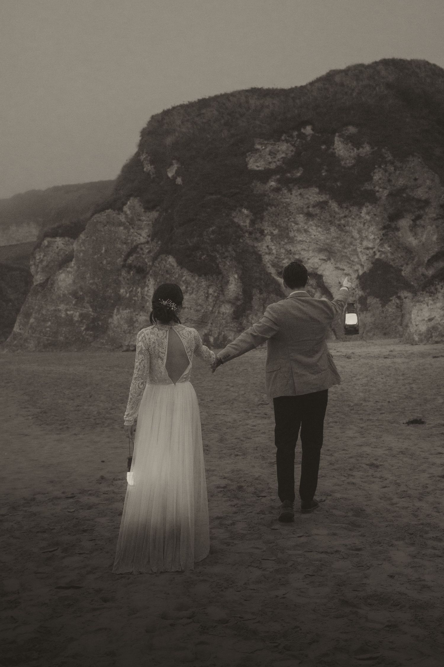 Couple walking along beach holding lanterns with dramatic cliffs in background.