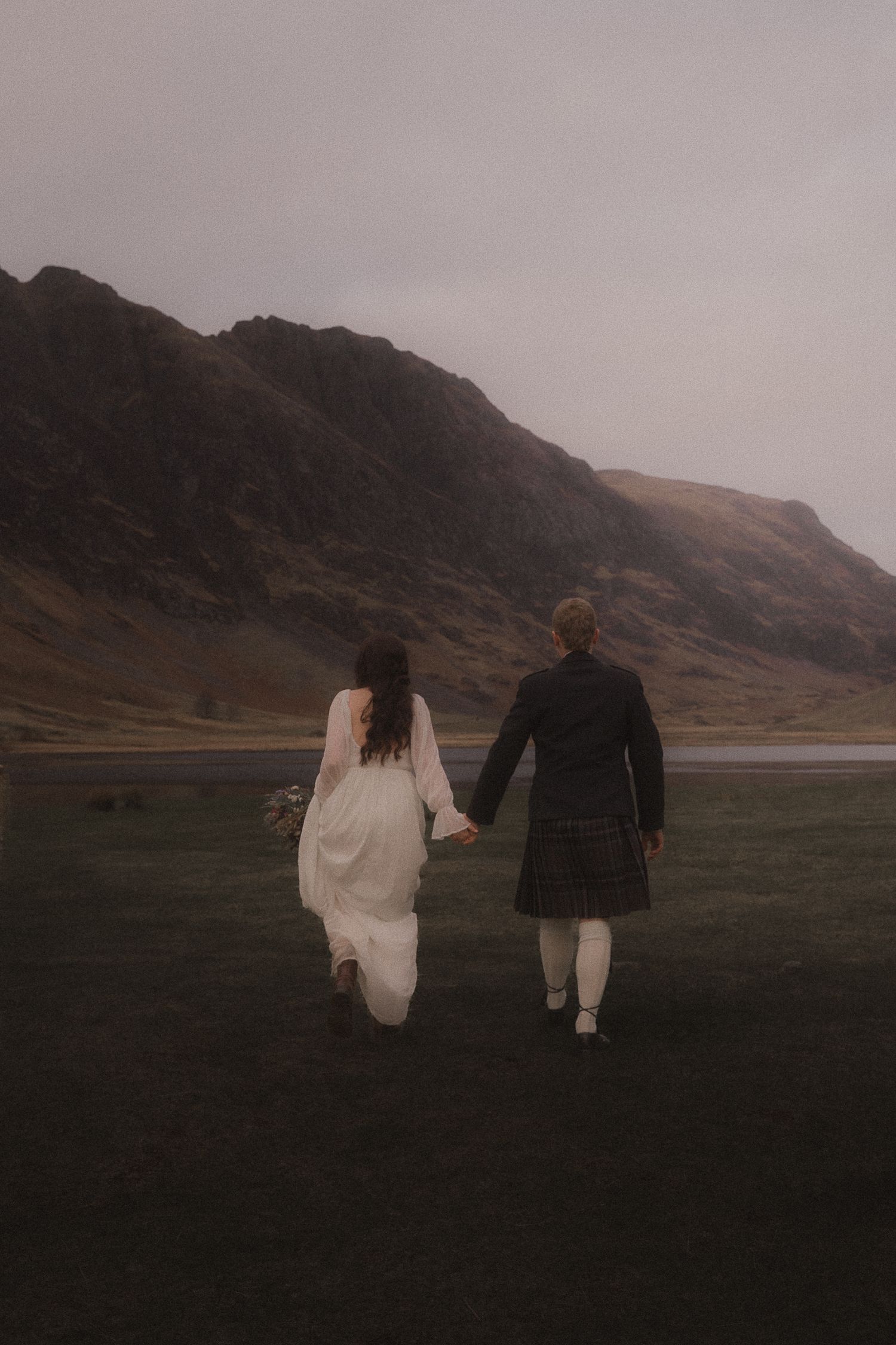 A couple holding hands walking together in traditional Scottish attire by a misty mountain loch in moody lighting.