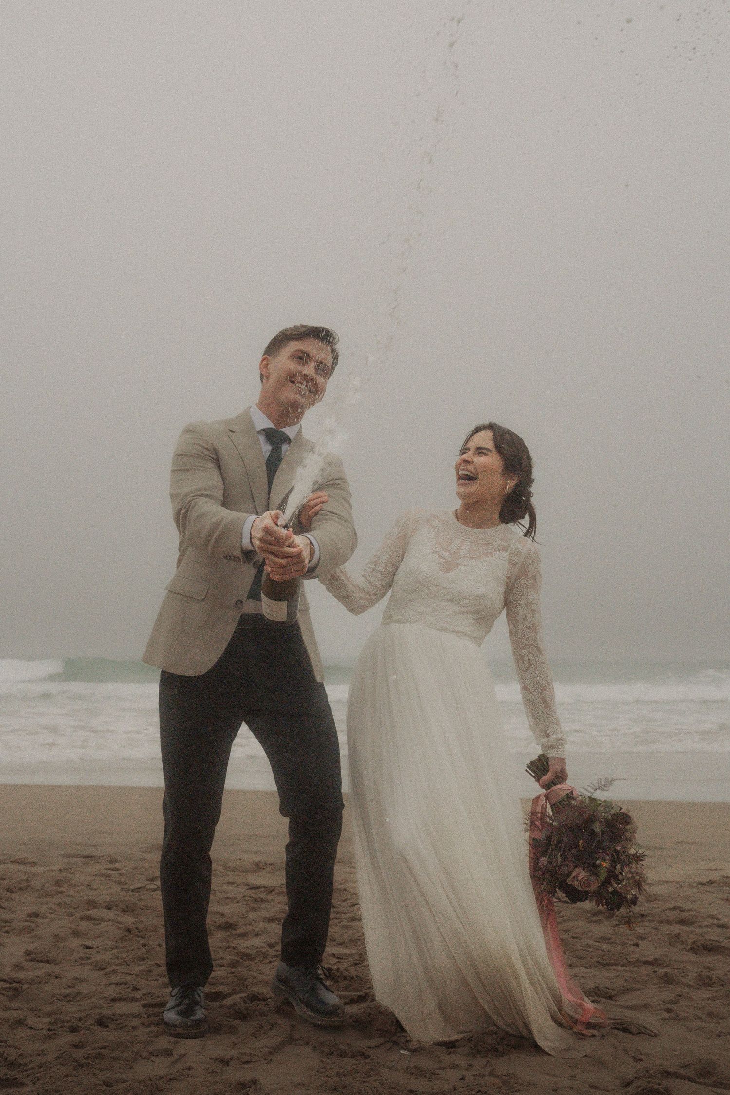 A joyful wedding couple celebrates with champagne on a misty beach.