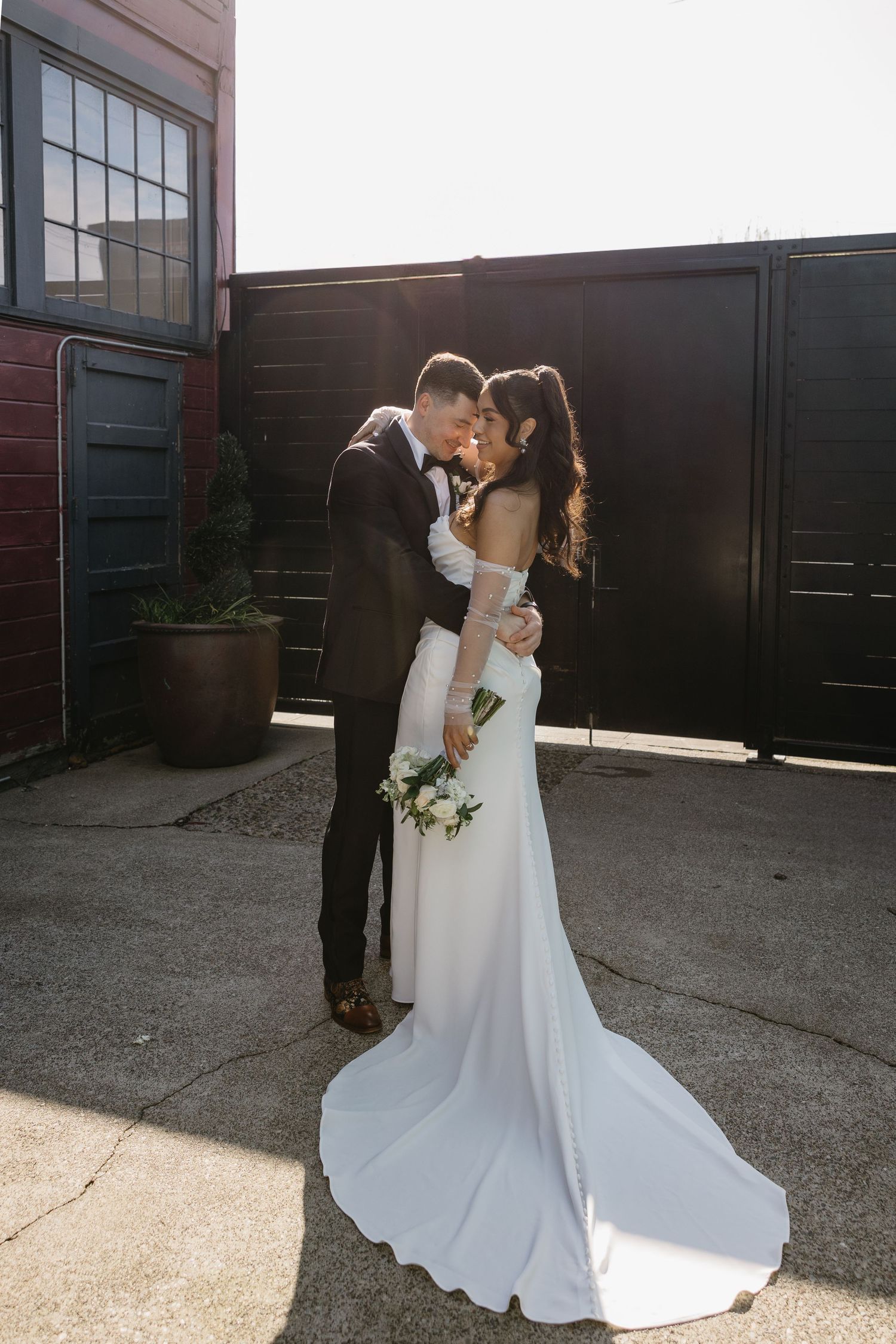 A couple in wedding attire shares an intimate moment against an industrial backdrop during golden hour.