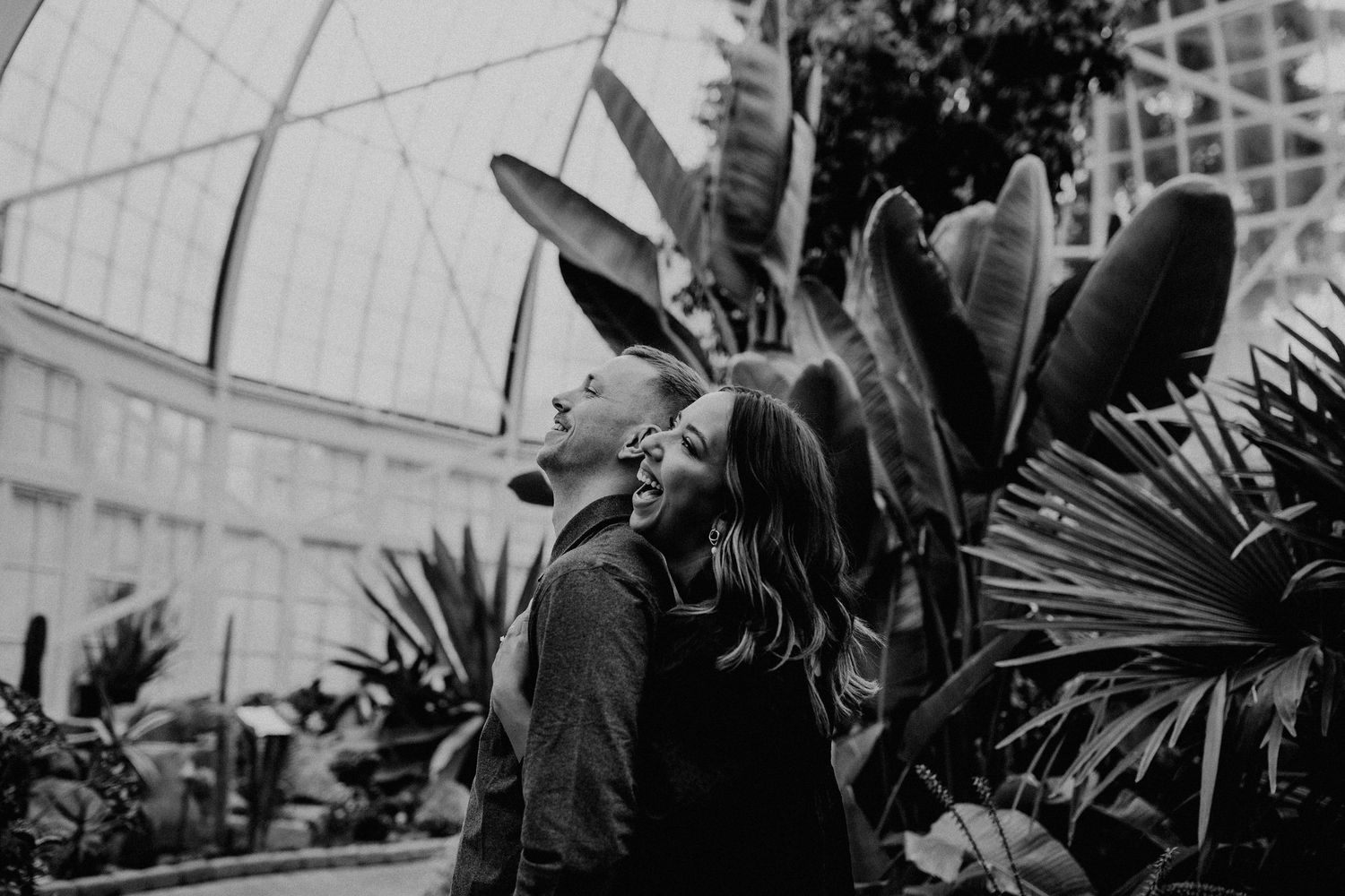 A black and white photo of two silhouettes standing near tropical plants inside a greenhouse setting.