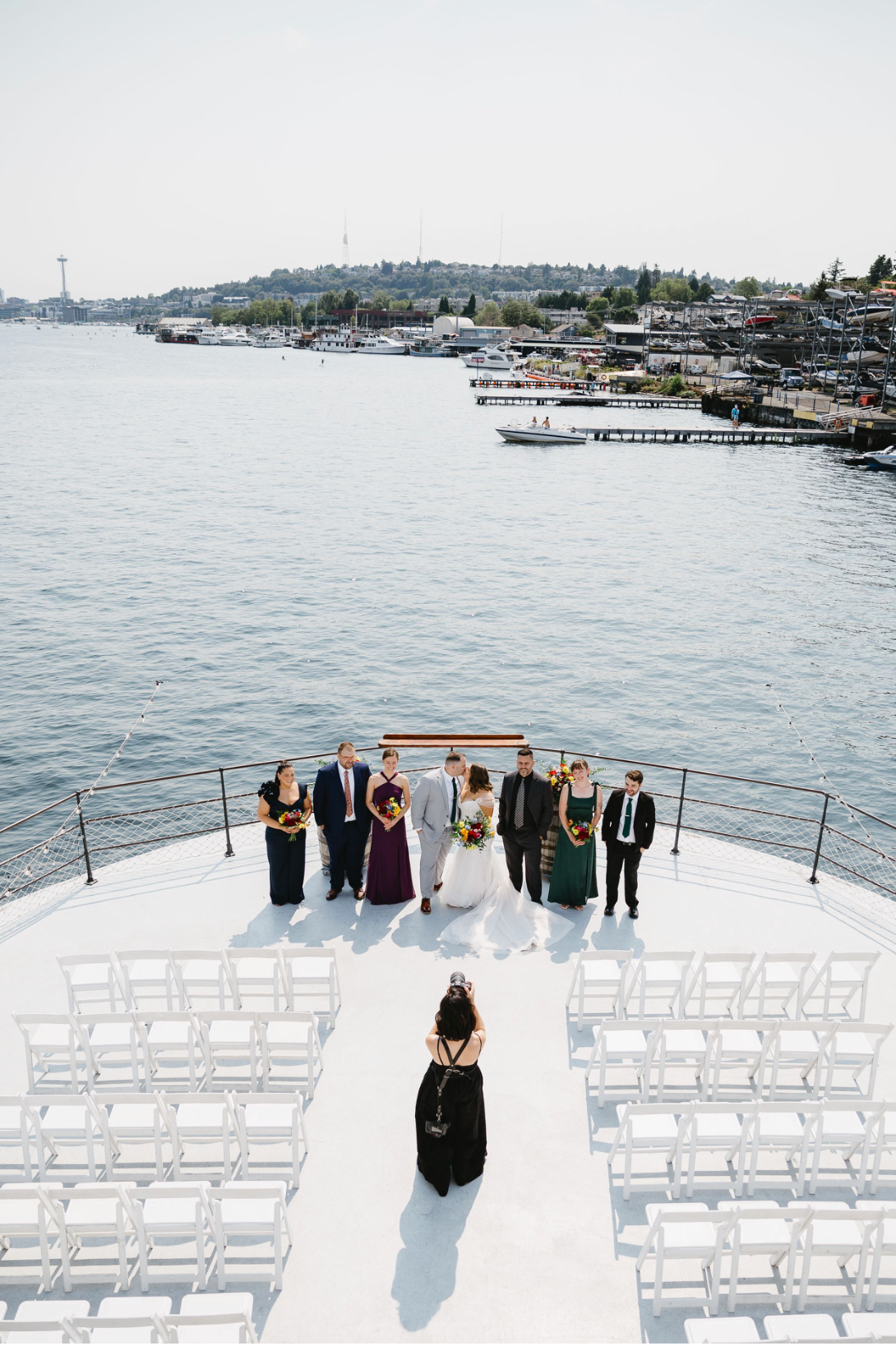 Wedding ceremony setup on yacht deck with white chairs and waterfront view of coastal homes and docks in background.