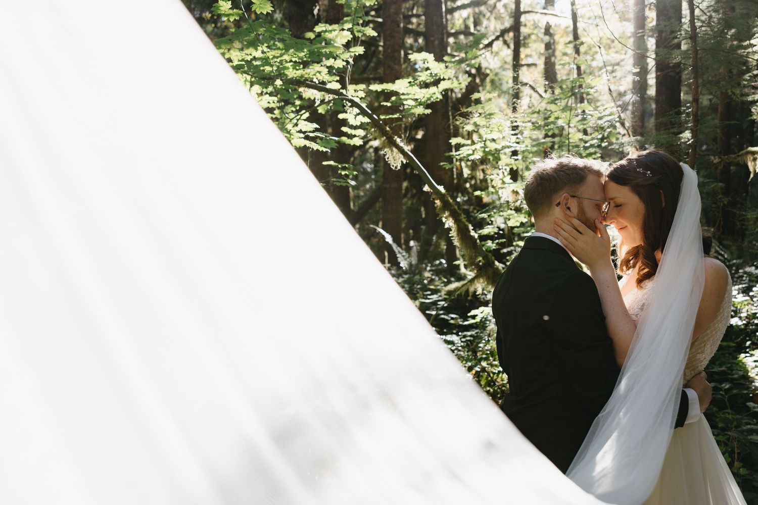 Wide angle shot of bride and groom embracing in sunlit forest with dramatic flowing white wedding dress.
