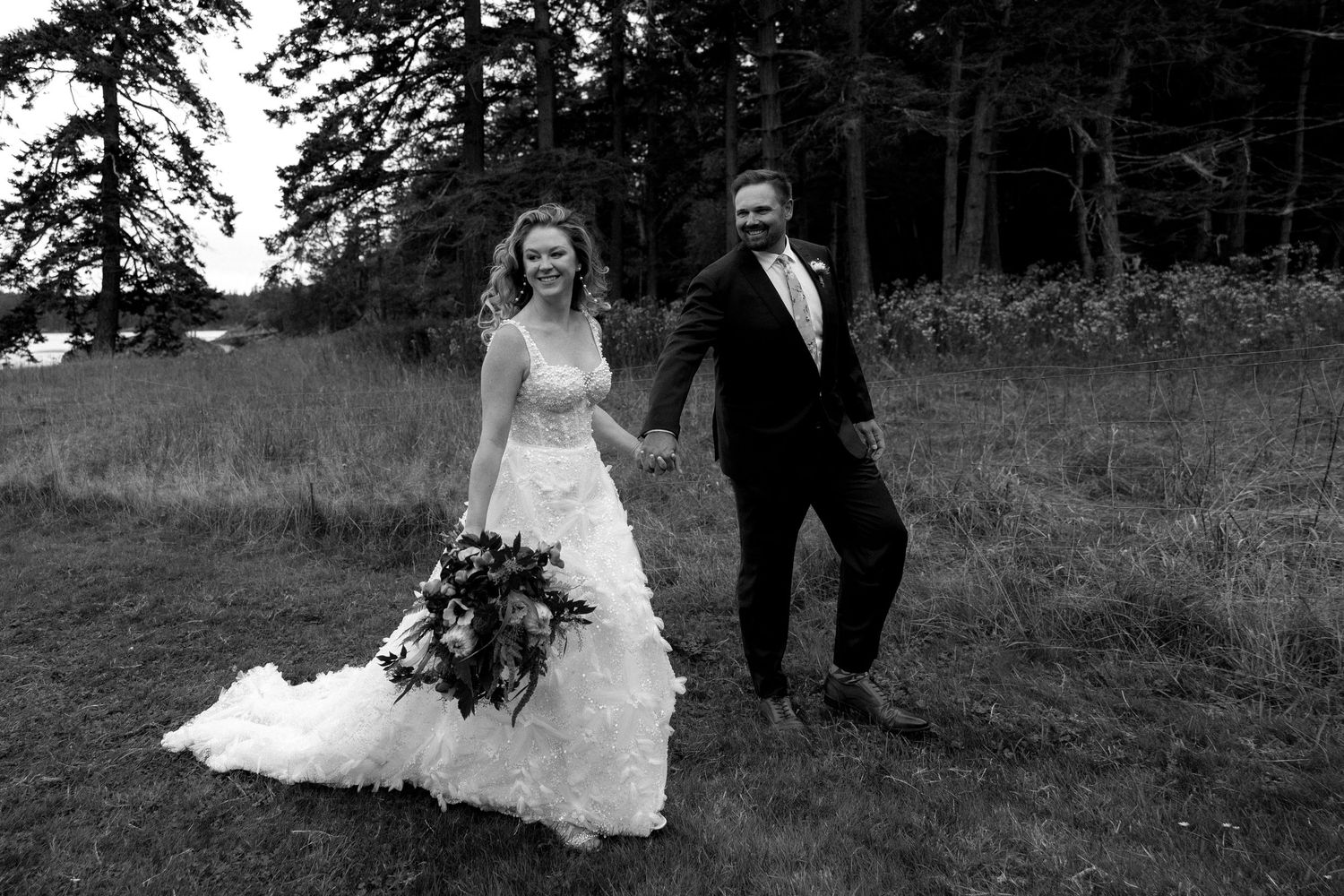 A newly married couple walks through a grassy field with evergreen trees in the background on their wedding day.