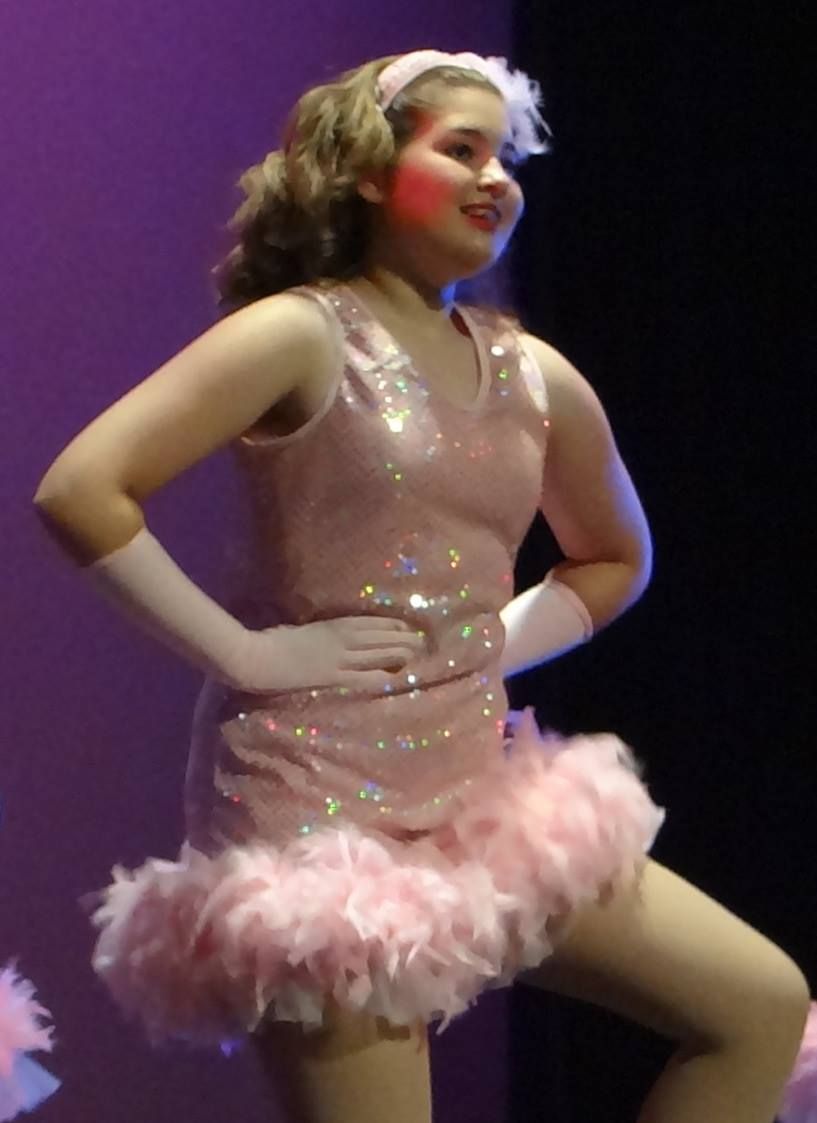 Performer in sparkly pink dance costume with feather trim on stage with purple lighting.