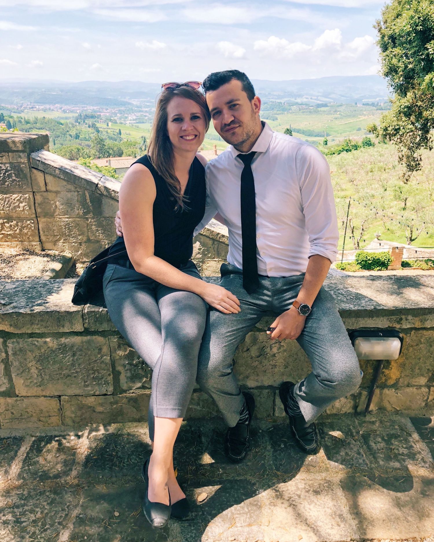 Casual couple sitting on stone wall with scenic mountain landscape behind them.