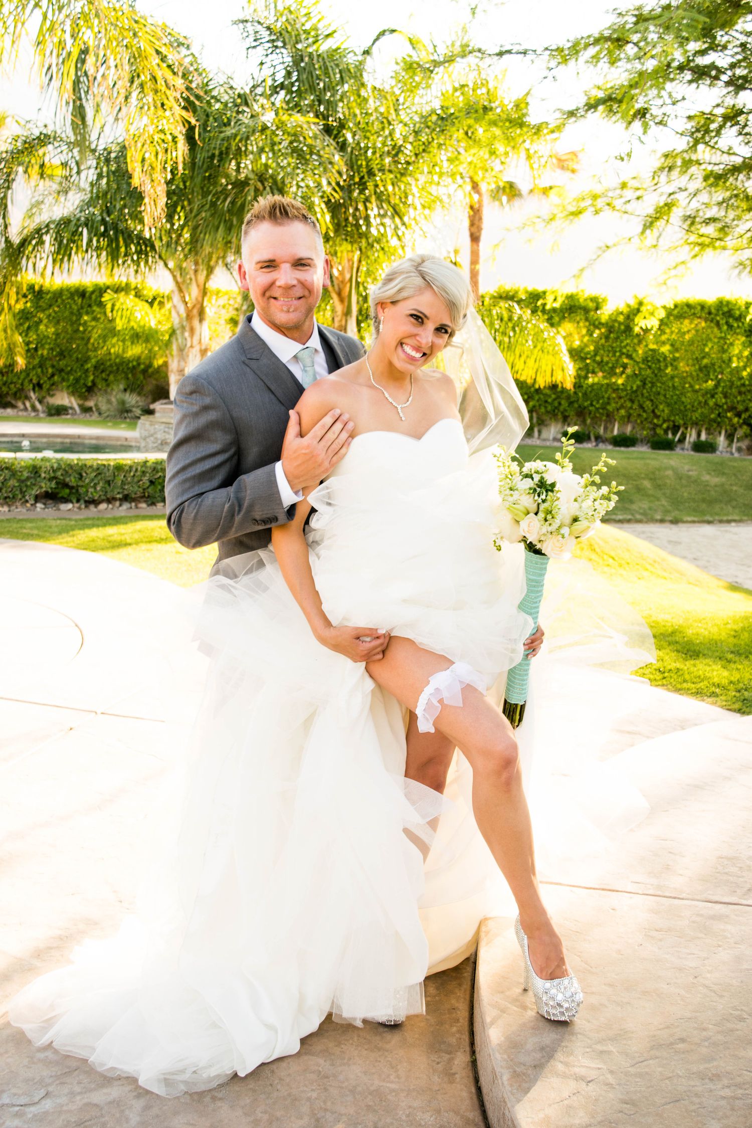 Playful wedding portrait in lush garden setting with palm trees and golden sunlight.
