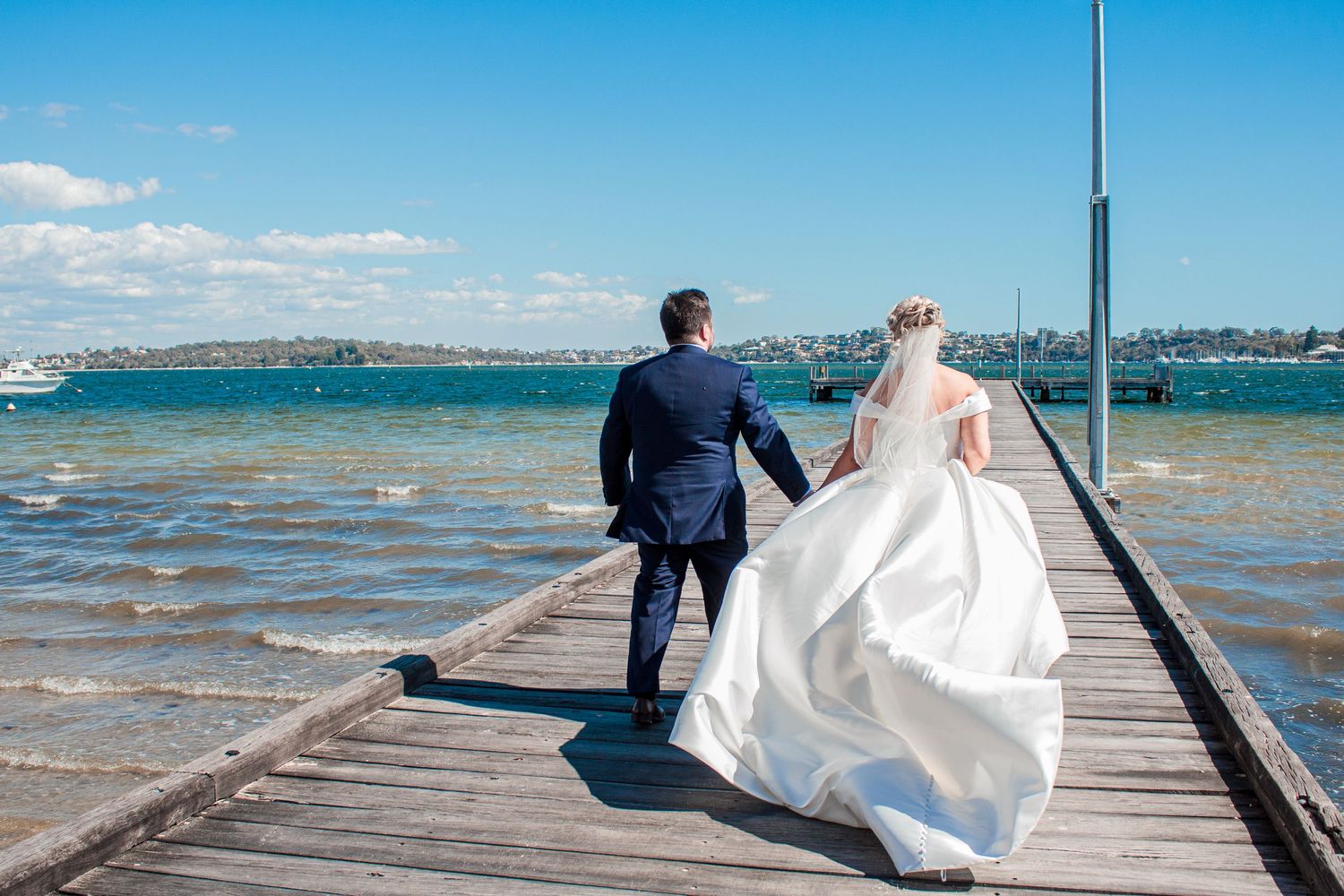 Couple walking on wooden pier by ocean in wedding attire with cityscape background.