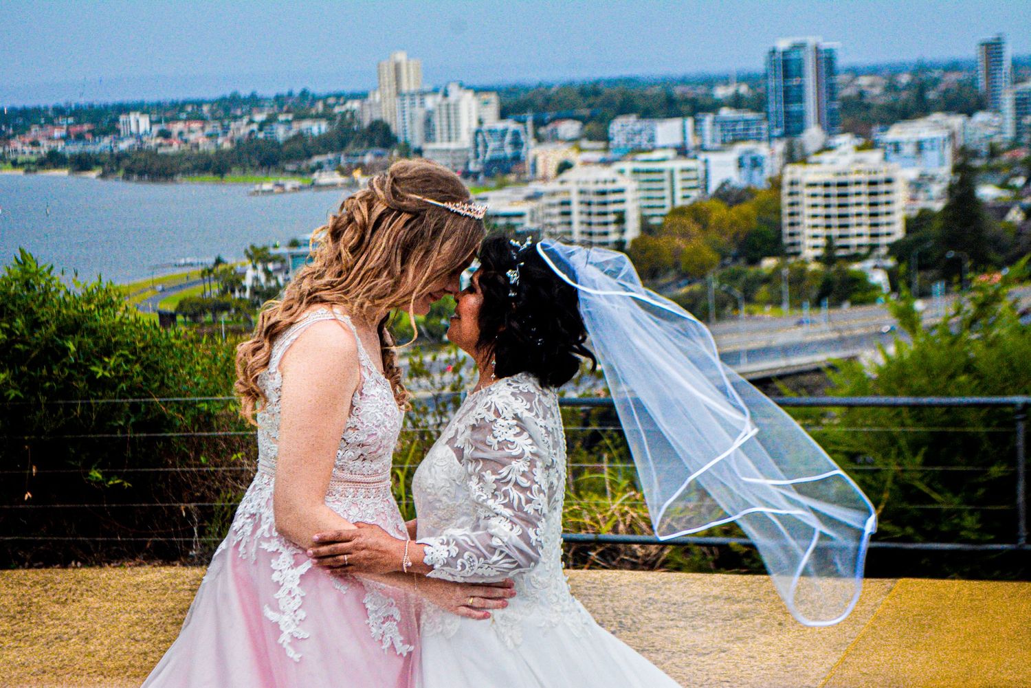 Wedding couple shares a romantic moment on balcony overlooking coastal cityscape with high-rise buildings.