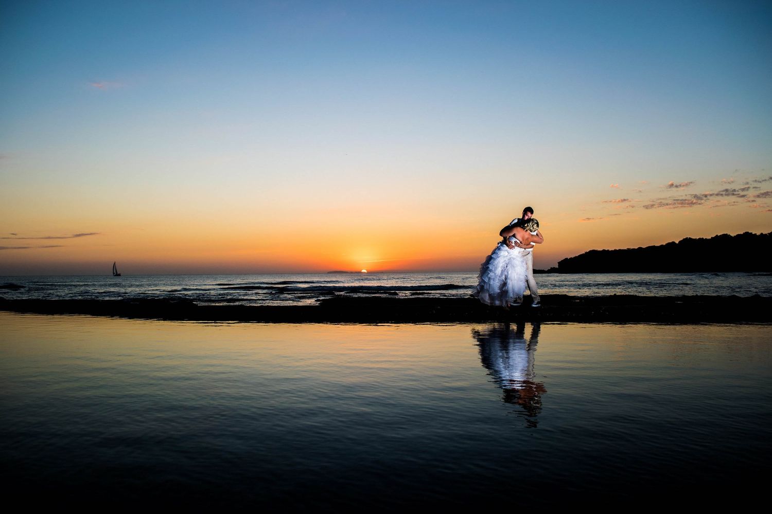 Silhouetted couple reflects in wet beach sand during dramatic ocean sunset.