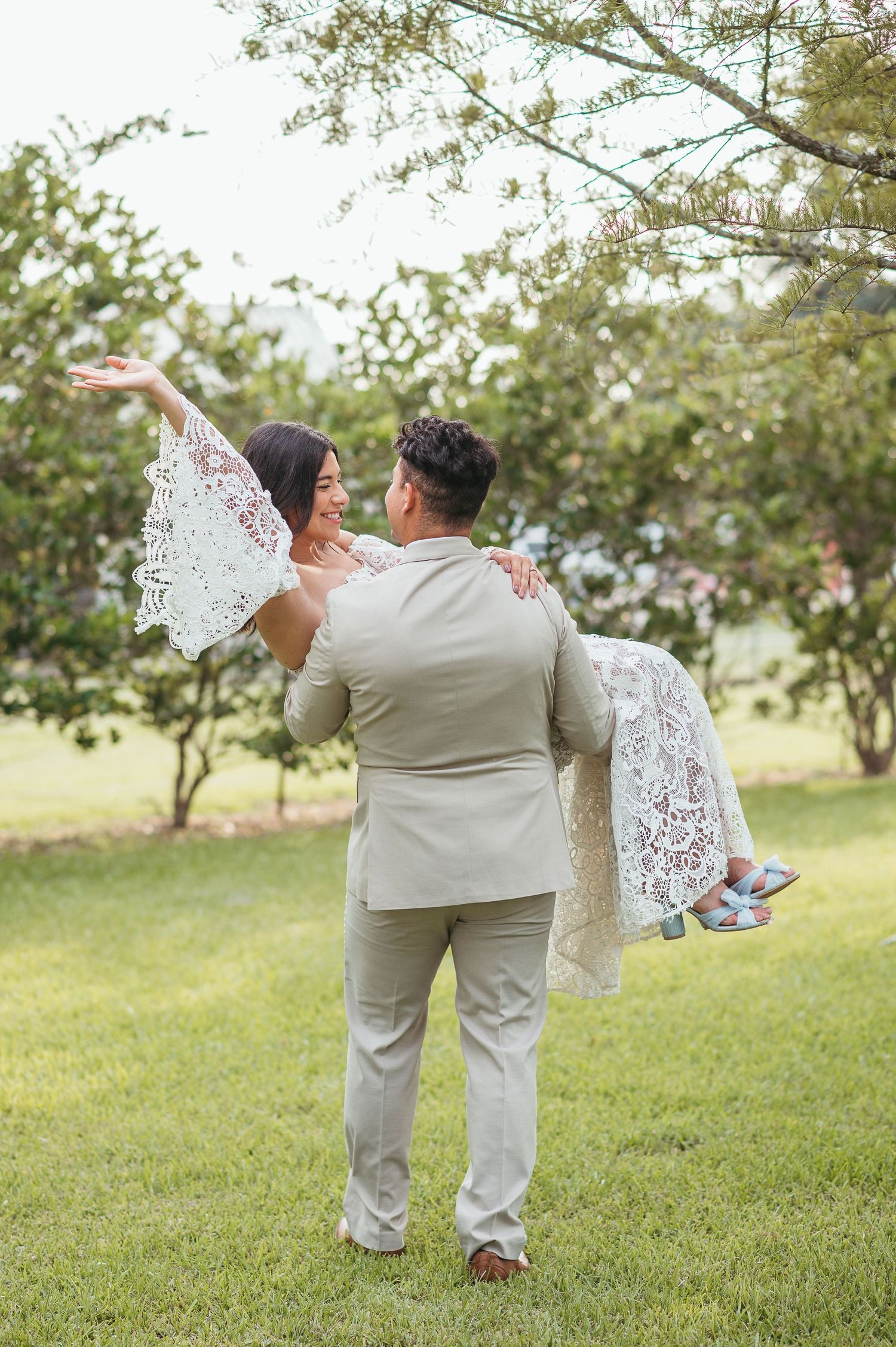 A romantic wedding photo of a groom carrying his bride in a lace dress through a garden setting.