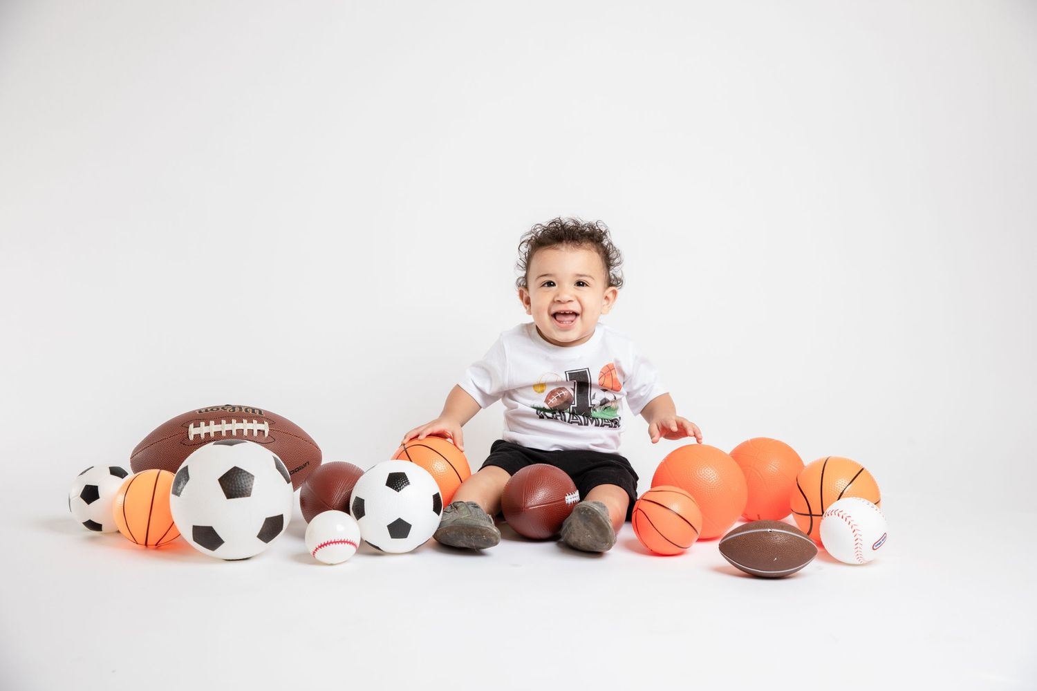 Happy toddler sits surrounded by various sports balls and plush toys on white background.