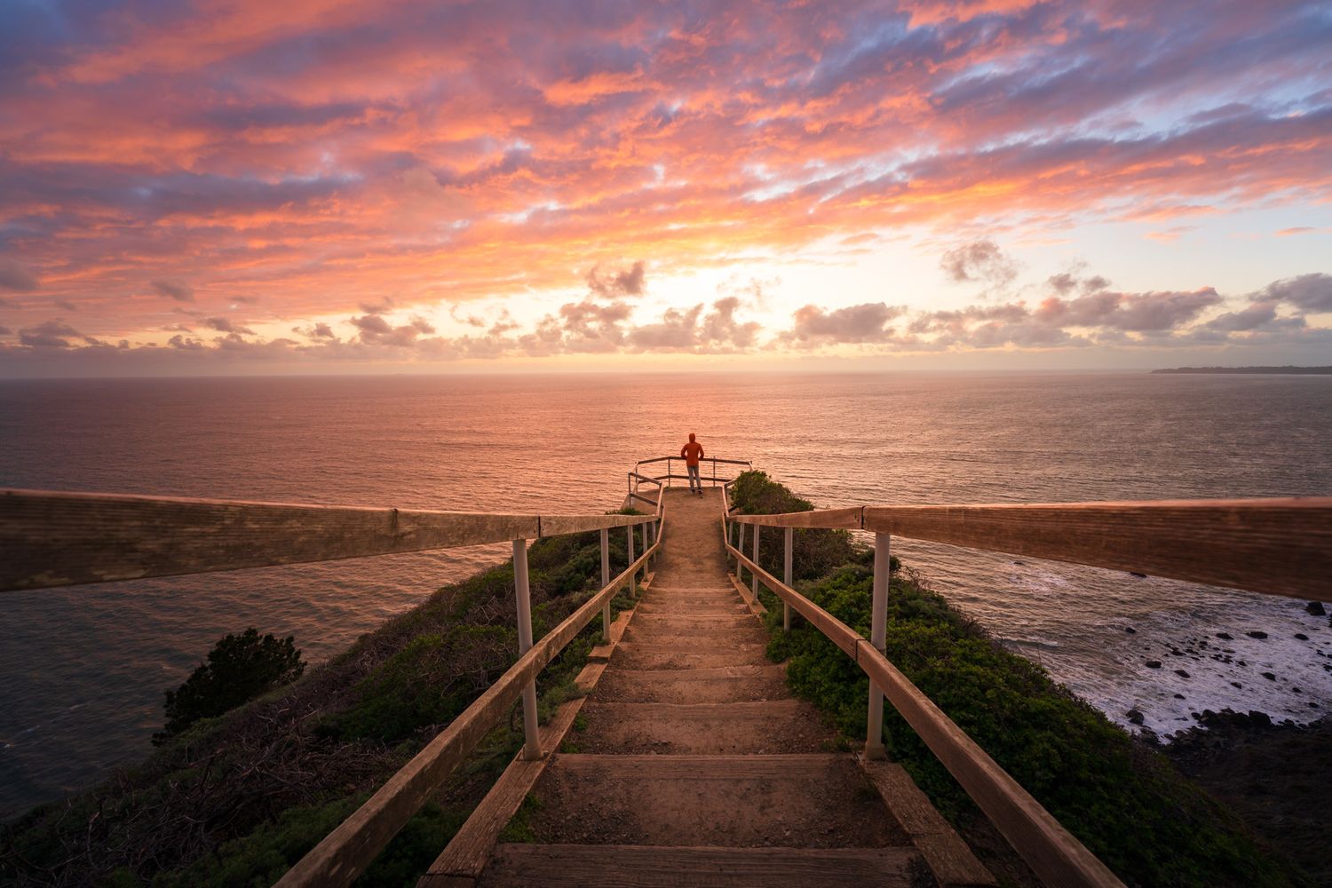 Wooden stairs descend toward the ocean under a vibrant sunset with pink and orange clouds.