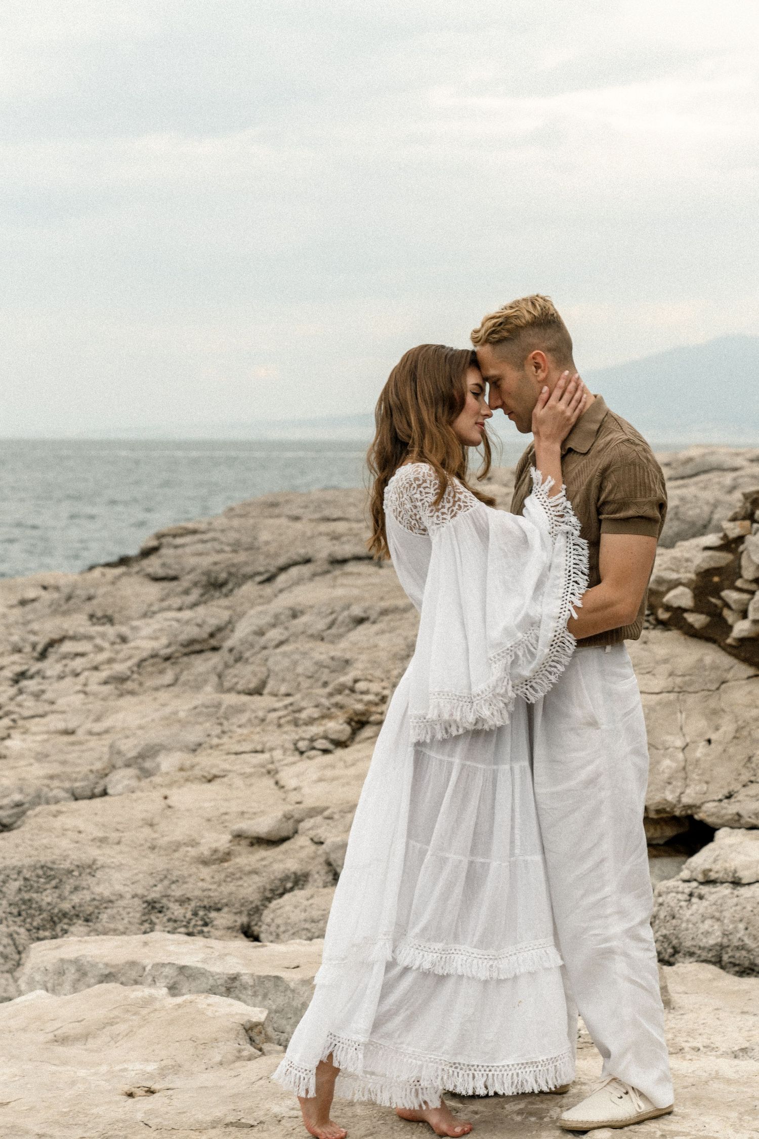 A romantic couple in white outfits embraces on rocky coastal cliffs against an ocean backdrop.