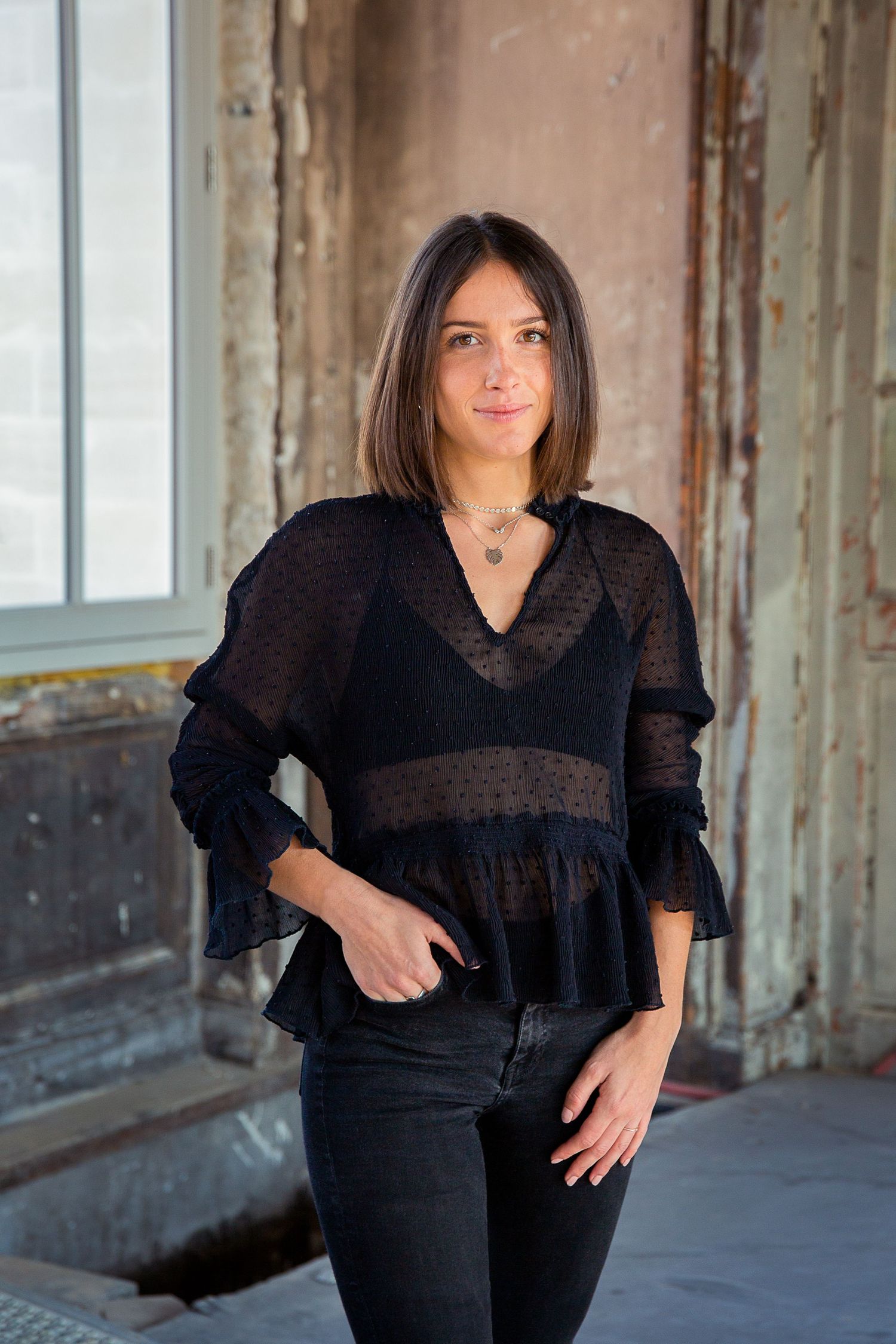 A fashion portrait of someone in a sheer black ruffled top and jeans against a rustic backdrop.