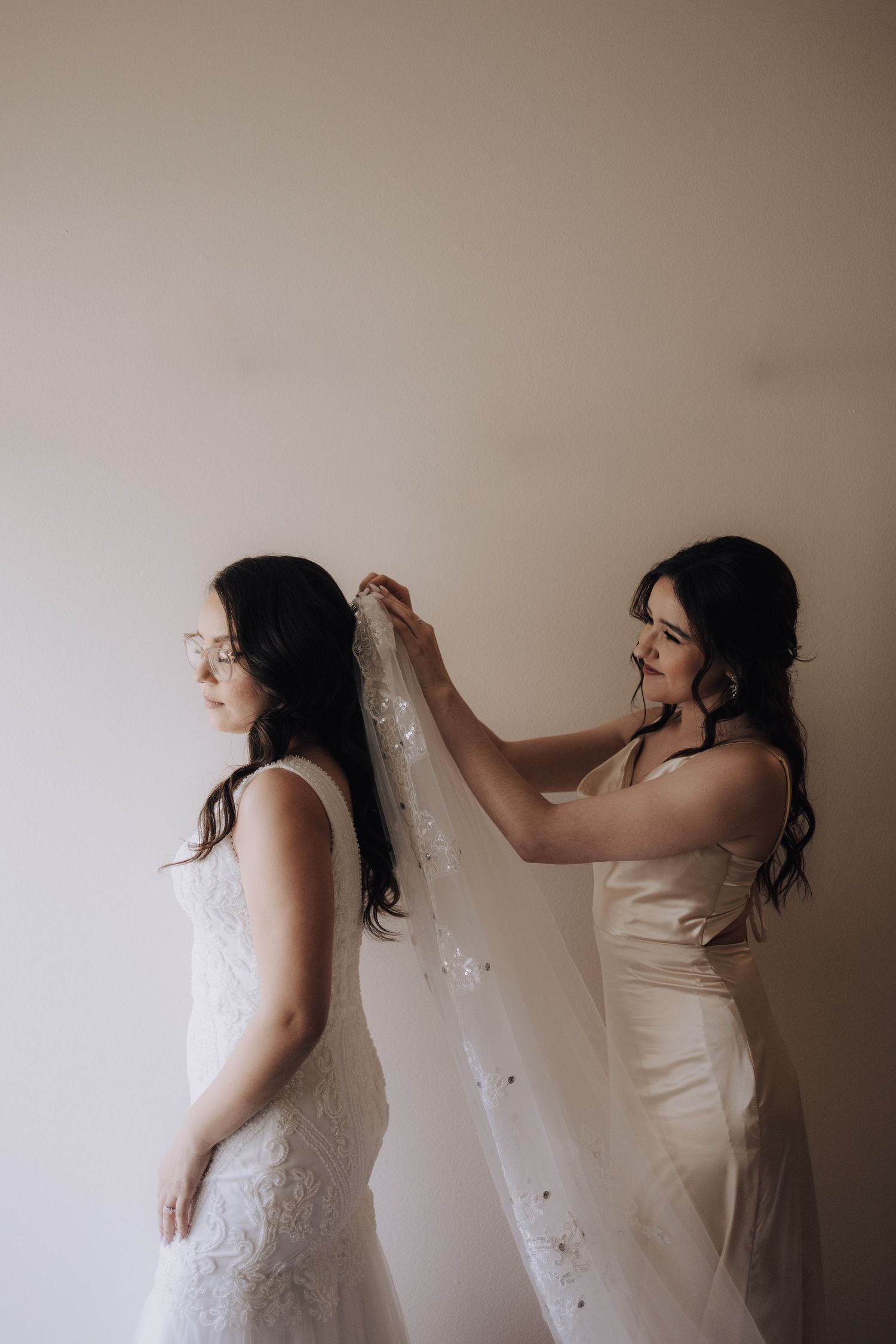 A bridesmaid helps adjust a wedding veil against a neutral wall background.