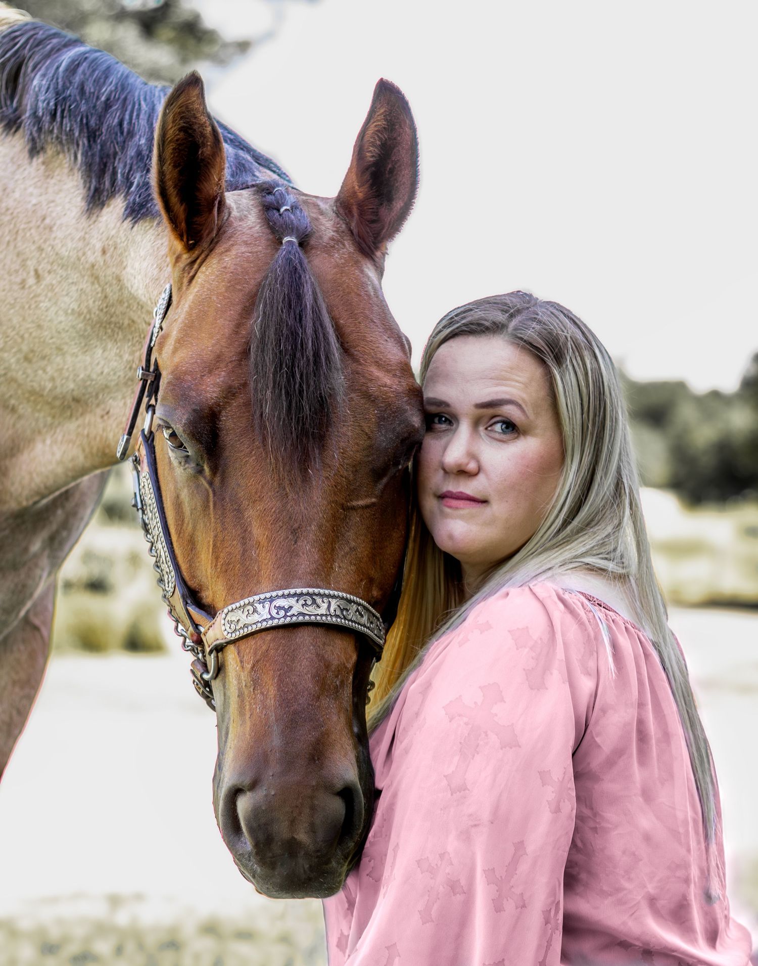 A blonde woman in a pink top leans affectionately against a chestnut horse wearing a decorative halter.