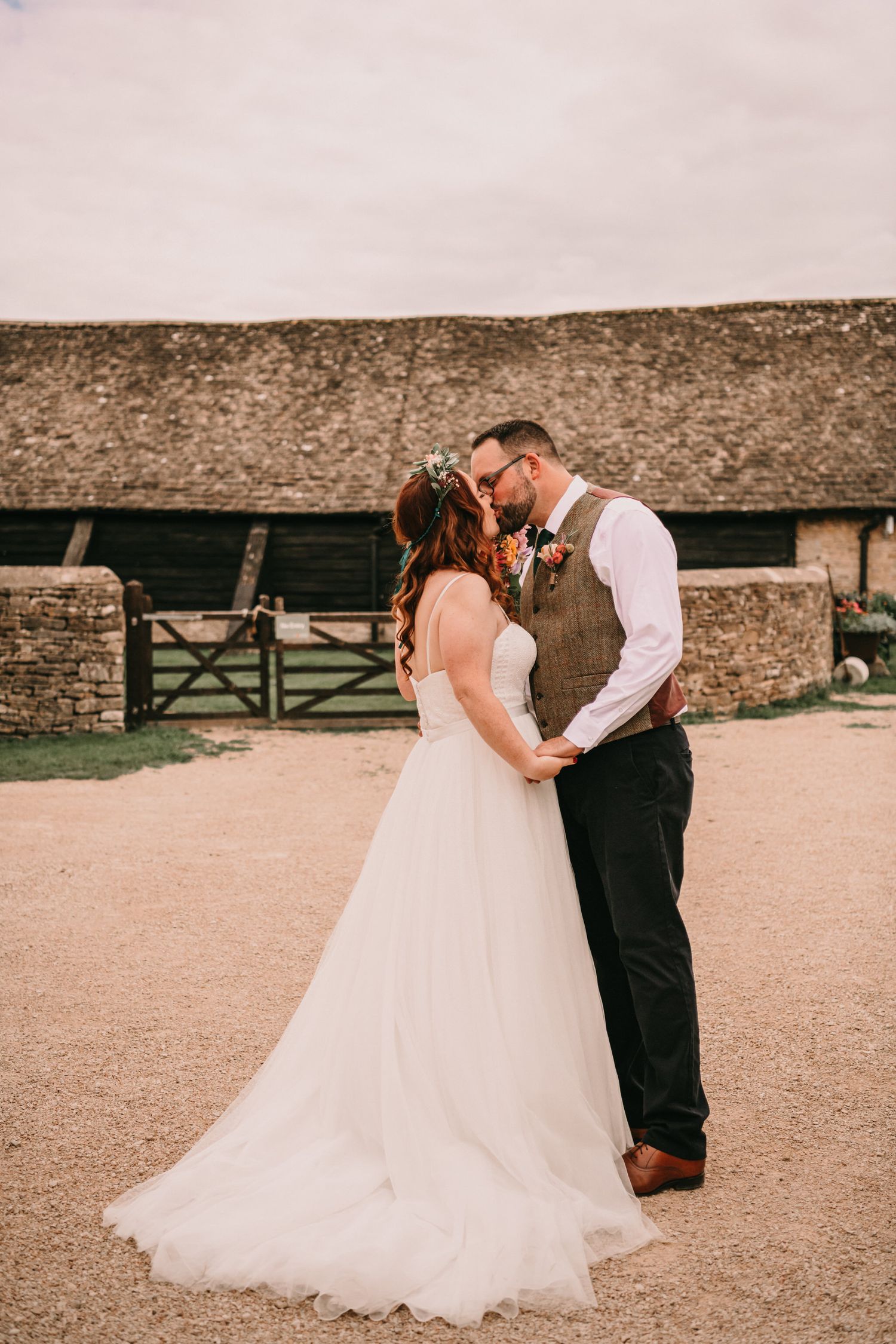 A romantic wedding couple sharing a kiss outside a rustic barn with stone walls and gates.