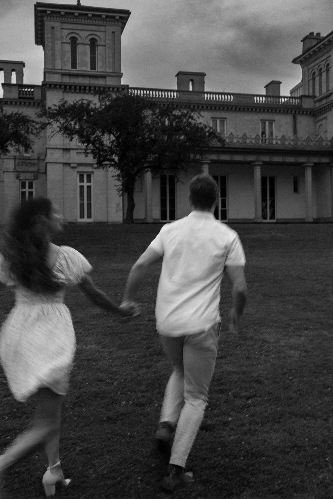 A blurred motion black and white photo of a couple running across a lawn in front of a historic mansion.
