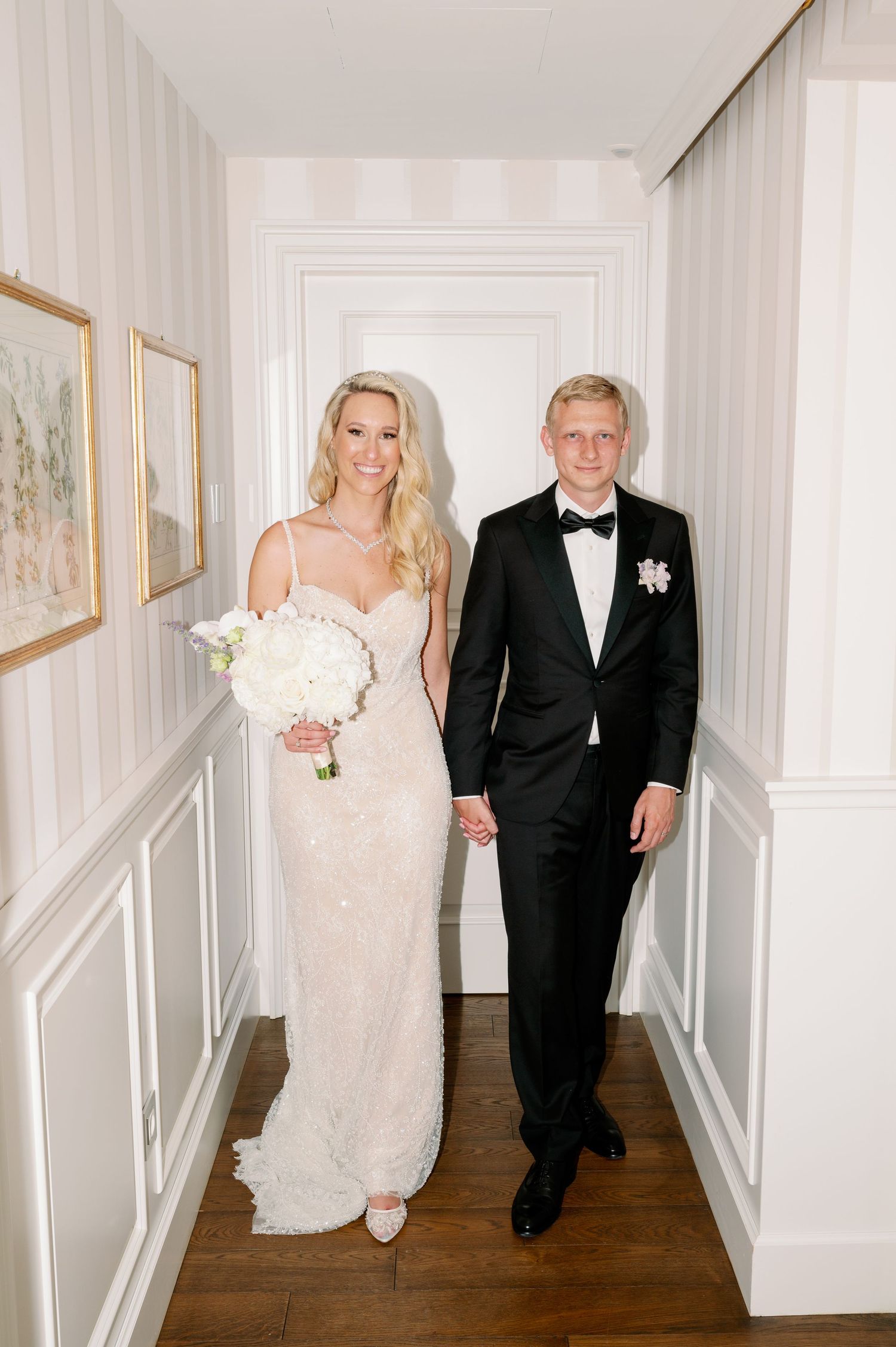 An elegant couple poses in a classic white hallway with wainscoting and artwork.