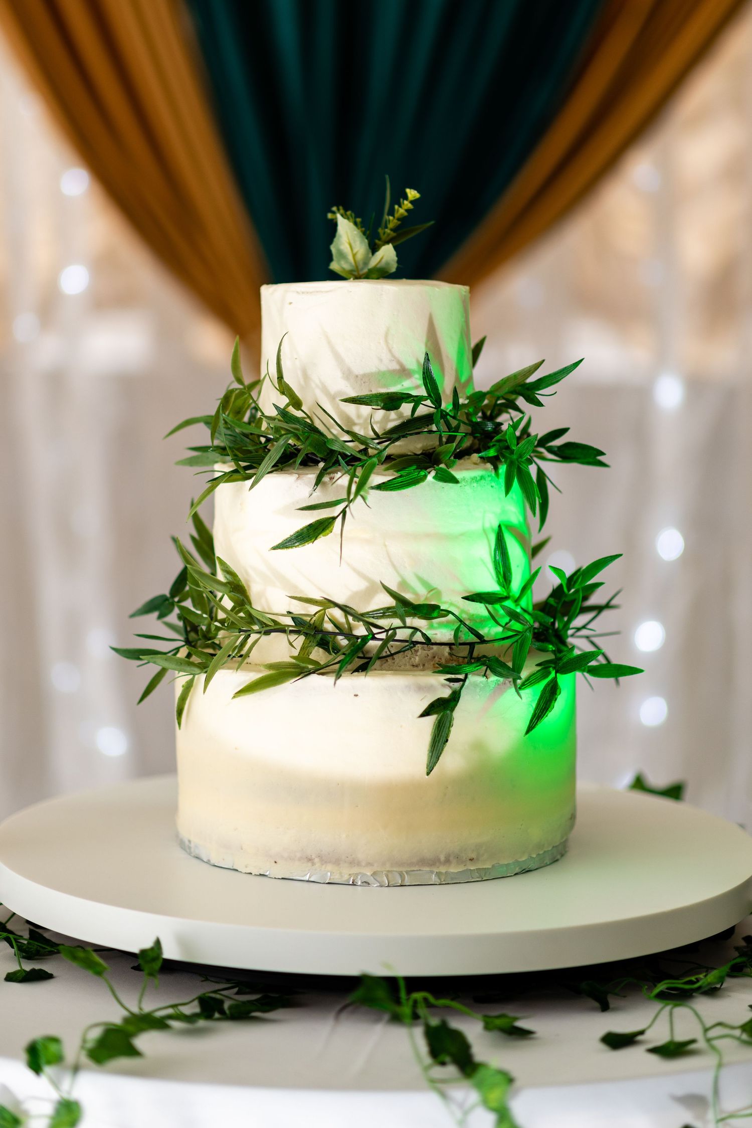 White wedding cake decorated with fresh greenery and leaves under soft lighting.