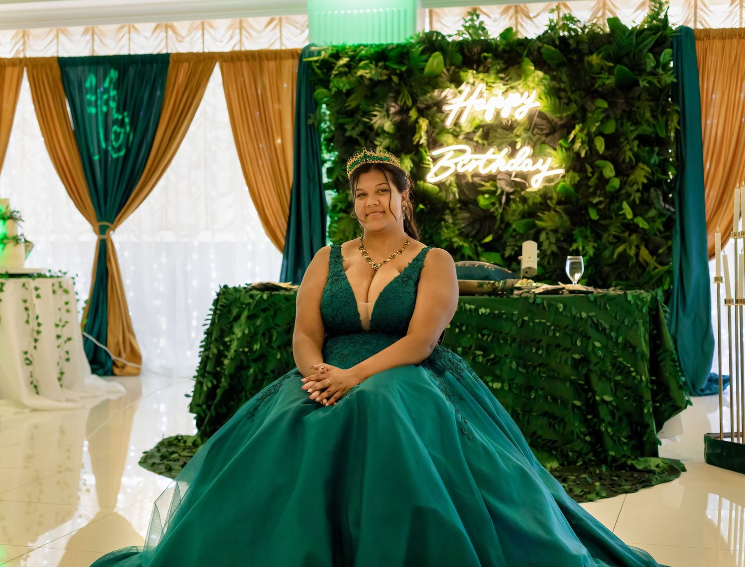 Wedding party members in emerald green dresses pose against a greenery wall backdrop.