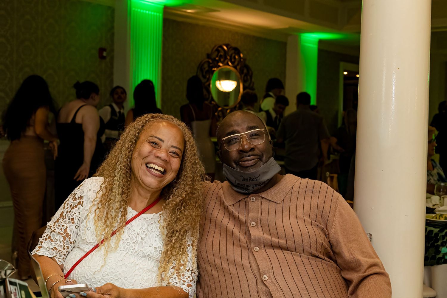 Party guests smiling and celebrating at venue with green ambient lighting and white columns.
