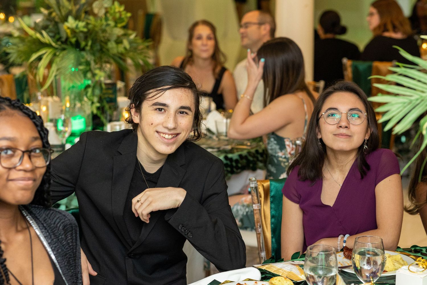 Guests enjoying dinner and conversation at formal event with green lighting and tropical plants.