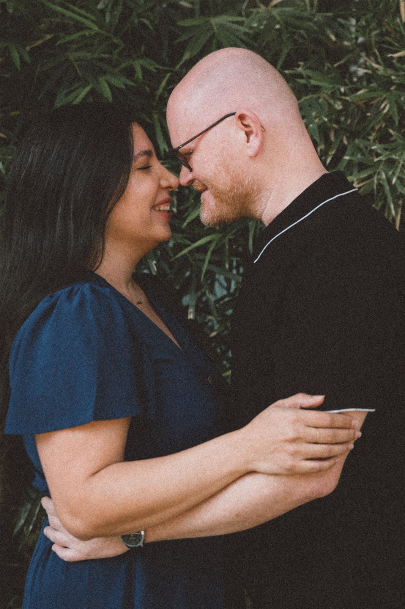 Intimate moment between couple in blue dress and black shirt against foliage.