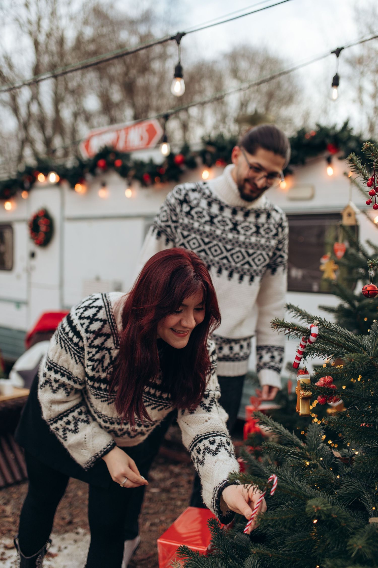 Couple in matching Nordic sweaters decorating Christmas tree outdoors with string lights.