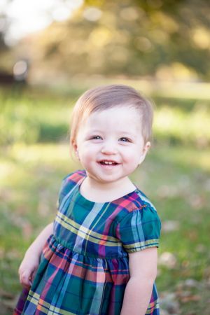 A happy baby in a plaid dress smiles brightly in an outdoor setting.