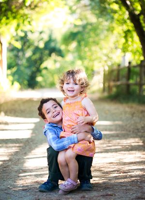 Siblings share joyful laughter while playing together on a sunlit forest path.