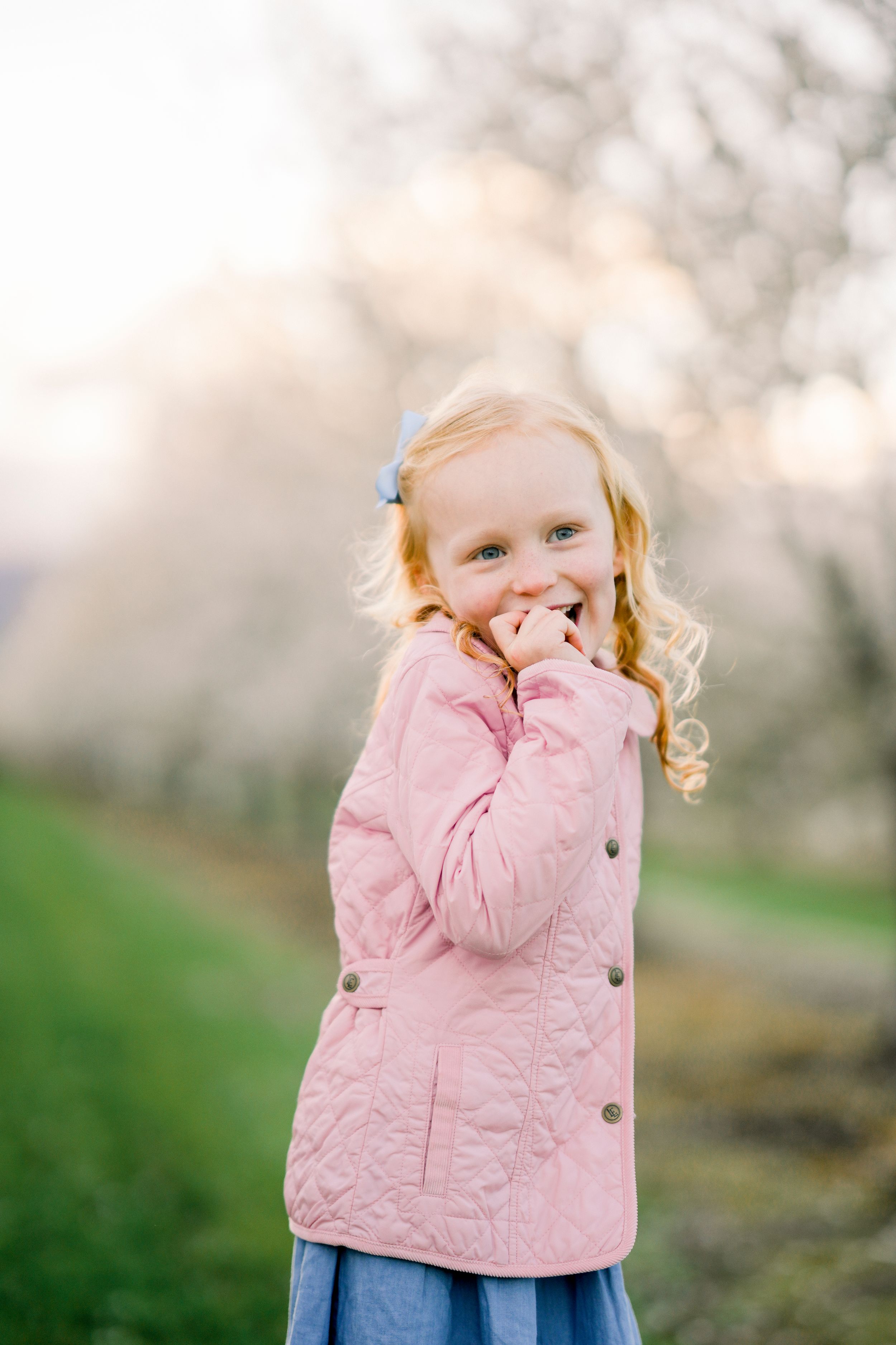 A young child in a pink quilted jacket poses playfully in a spring orchard setting.