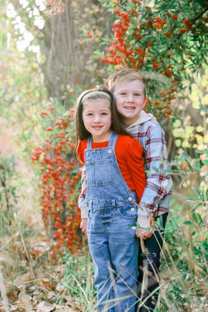 Two children in overalls and red shirts play together among red berries and green foliage outdoors.