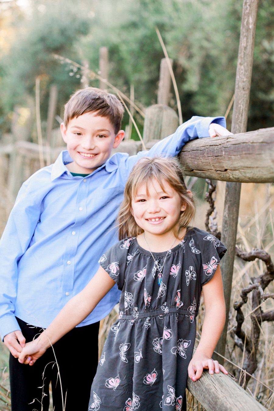 Two children pose together by a rustic wooden fence in casual clothing.