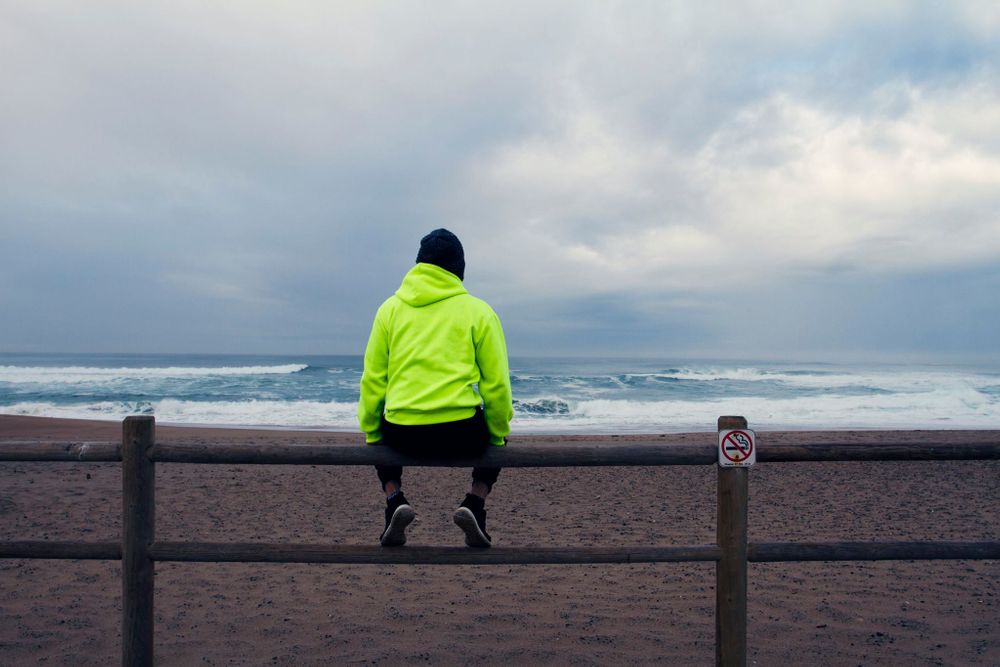 Person in neon yellow jacket sitting on railing overlooking stormy ocean.
